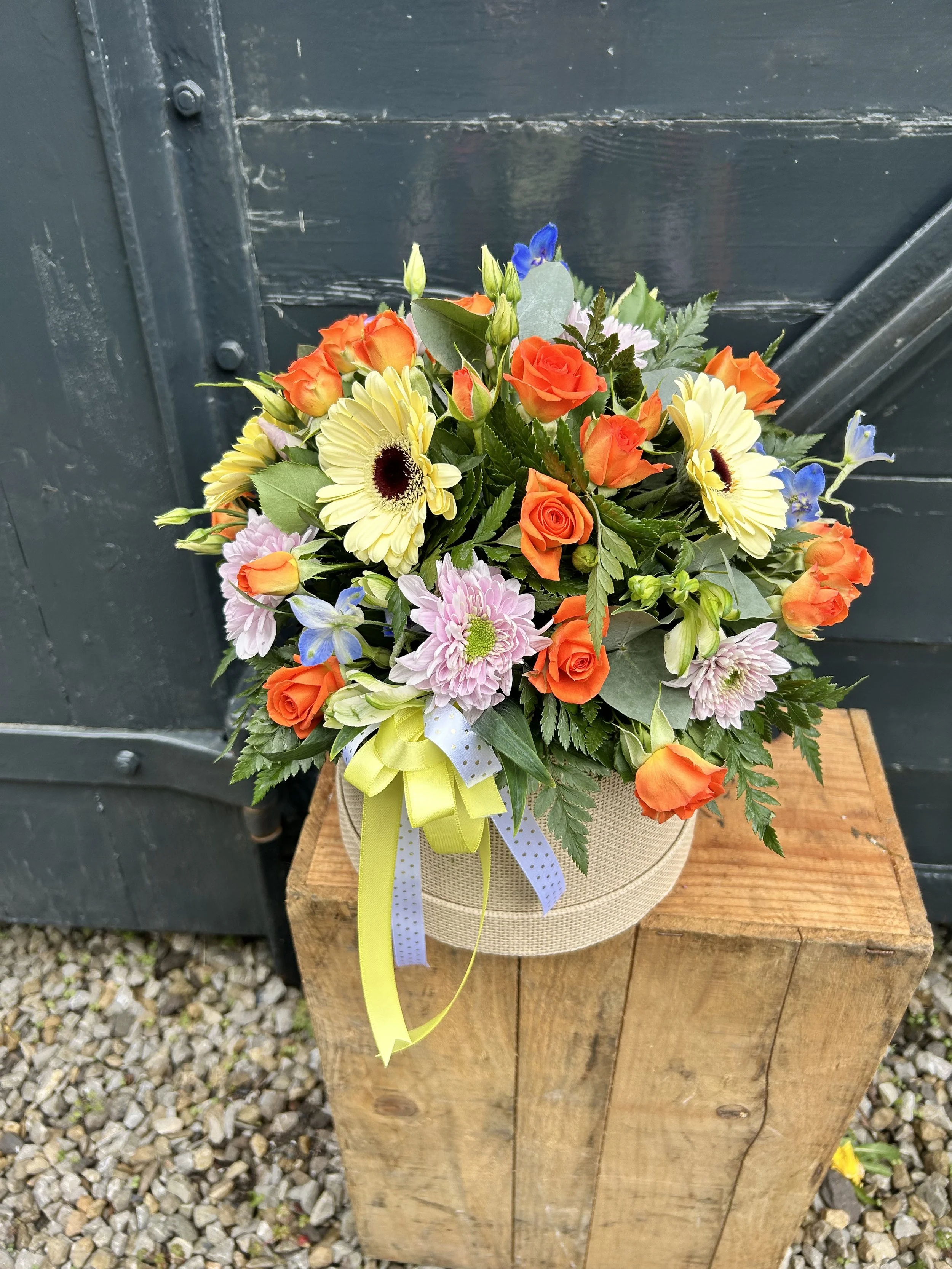 Colorful flower arrangement with orange roses, yellow gerbera daisies, pink chrysanthemums, blue delphiniums, and greenery in a beige hat box with yellow and white ribbons, placed on a wooden table outdoors.