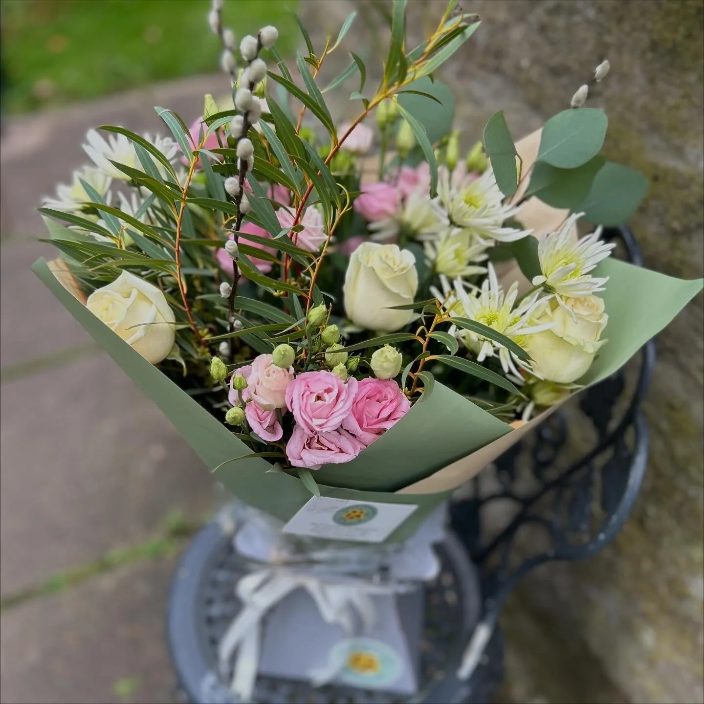 A bouquet of mixed pink, white, and yellow flowers with green leaves wrapped in green paper, placed on a decorative black metal table outside.