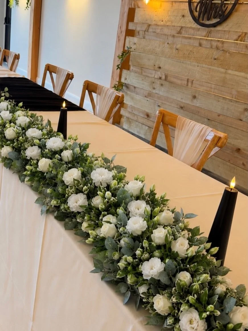 A wedding table decorated with a long bouquet of white flowers and greenery.
