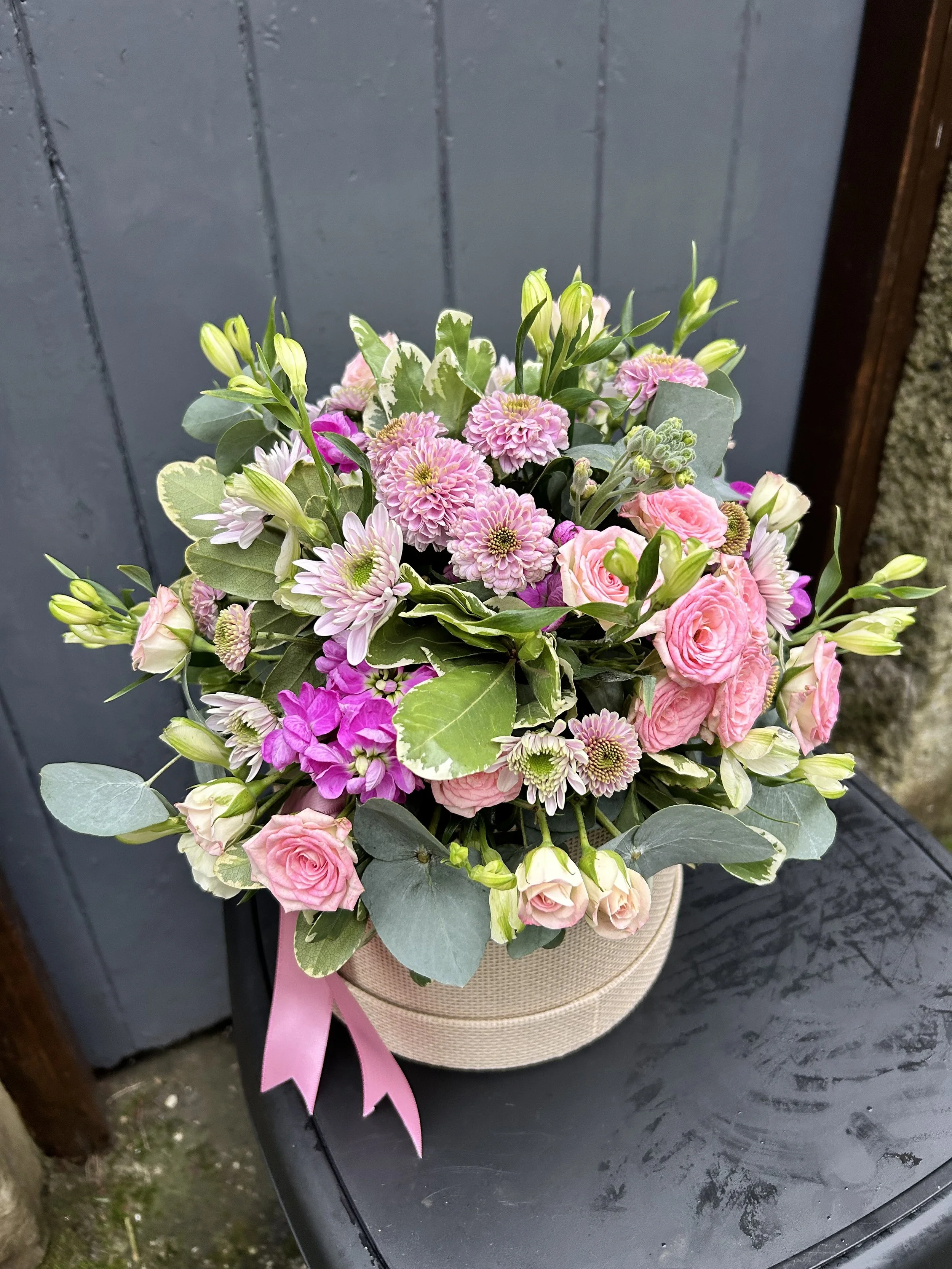A round beige flower bouquet with pink roses, pink chrysanthemums, white daisies, and green foliage, tied with a pink ribbon, placed on a black surface outdoors.
