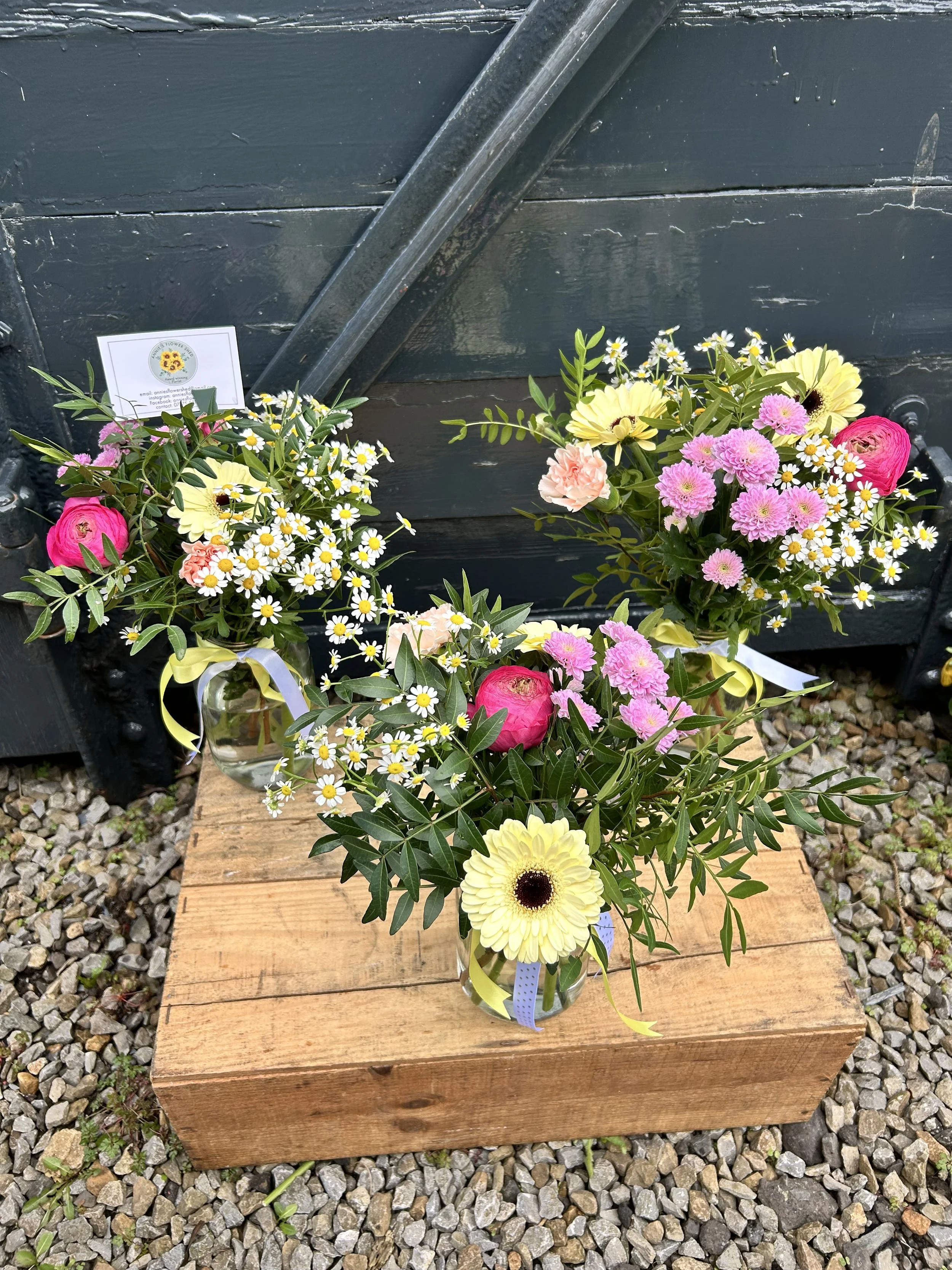 Flower arrangement with pink, yellow, and white flowers on a wooden block, with a black wall and gravel ground in the background.