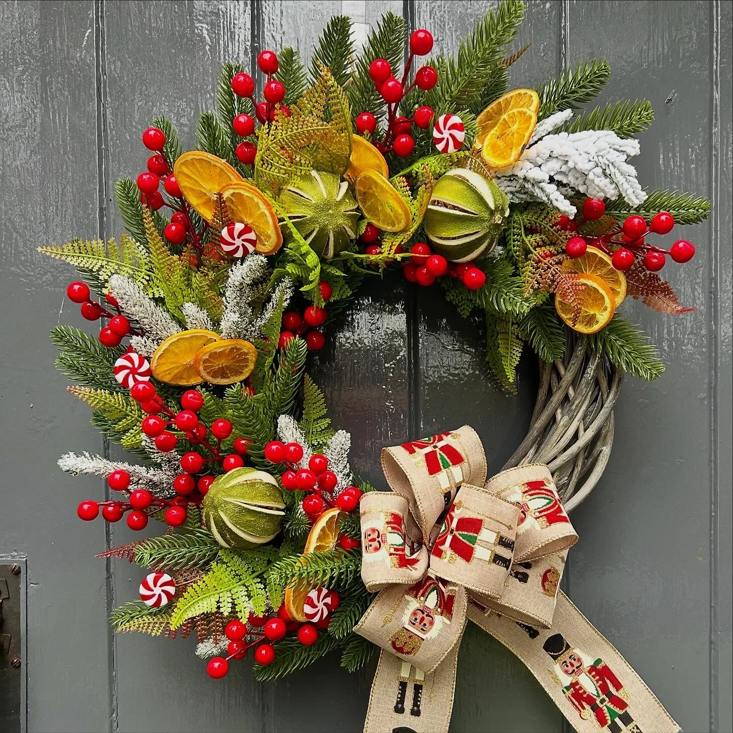 Christmas wreath with pine branches, red berries, dried orange slices, peppermint candies, frosted greenery, and a ribbon with nutcracker and holiday motifs hanging on a gray wooden door.