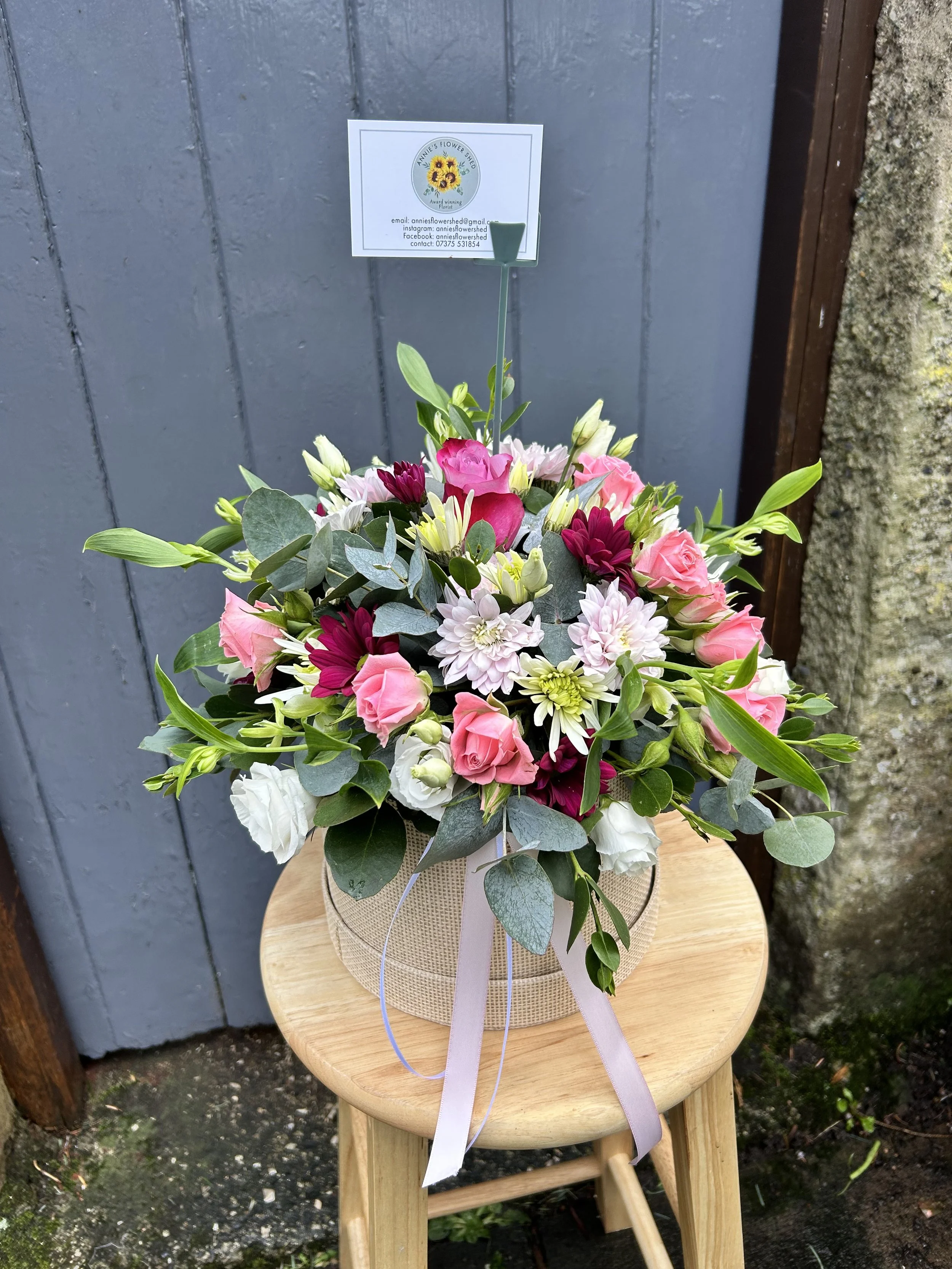A flower bouquet with pink roses, white lilies, purple chrysanthemums, and greenery arranged in a basket, placed on a wooden stool outdoors.