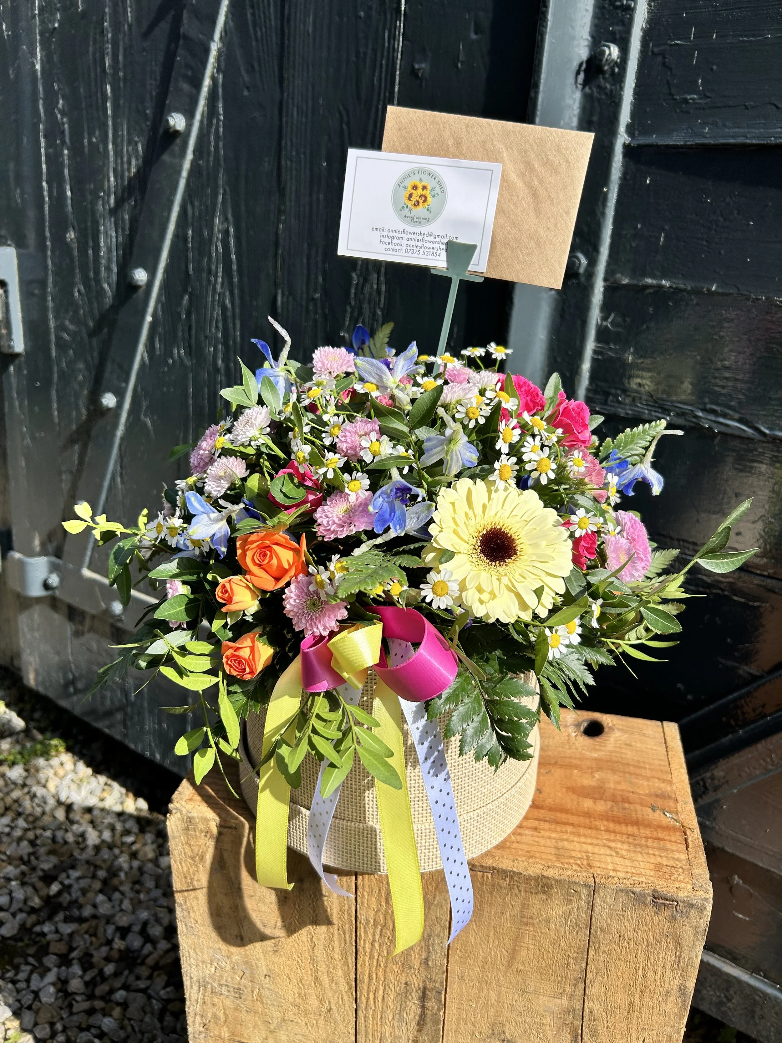 A colorful flower bouquet with pink, orange, yellow, blue, and white flowers and green leaves, wrapped in yellow netting and tied with pink, yellow, and white ribbons; placed on a wooden surface in front of a black wooden fence with a small sign show
