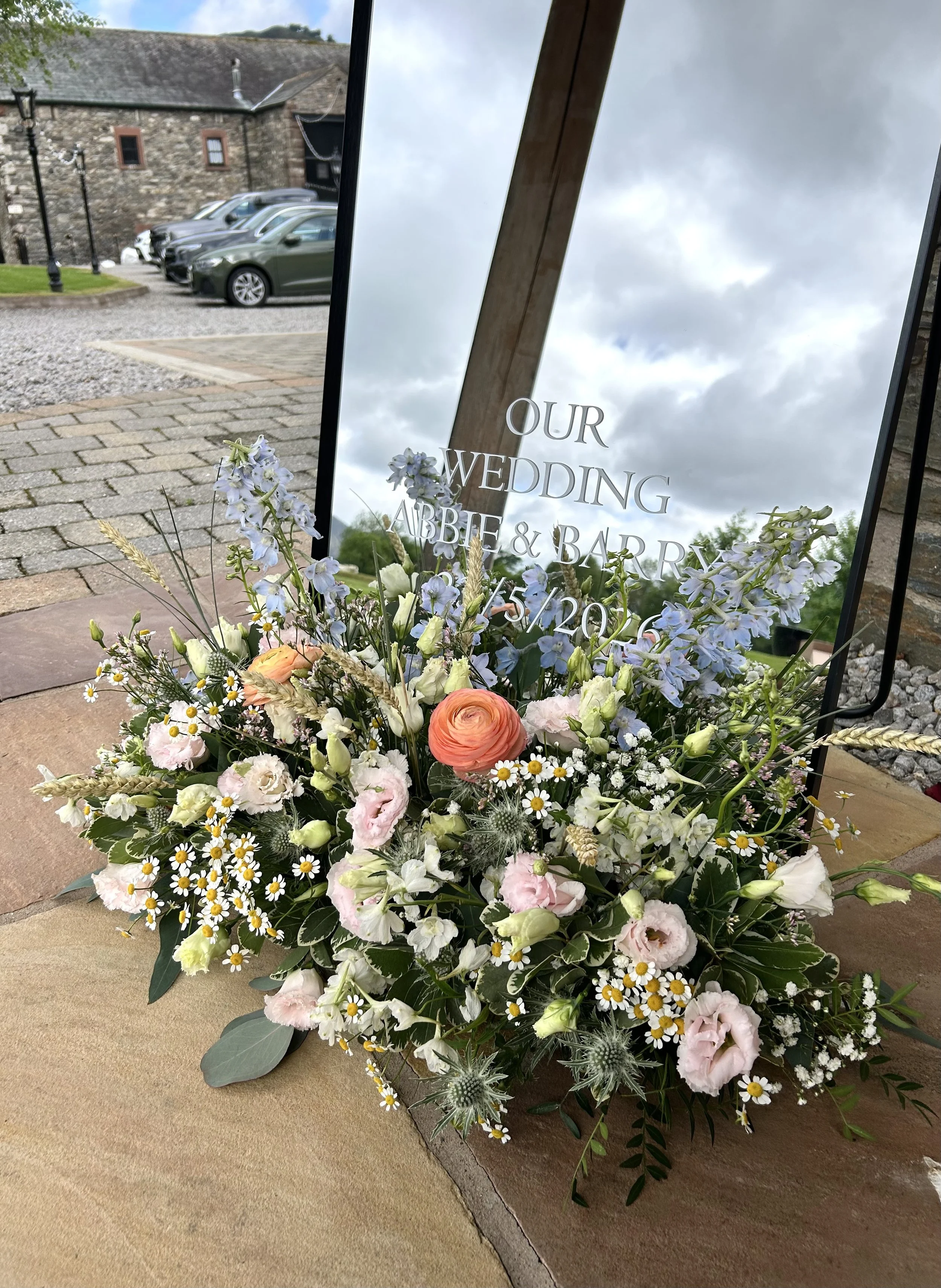 A colorful flower arrangement with various white, pink, peach, and purple flowers, complemented by green foliage, placed in front of a reflective sign for a wedding between Abbie and Barry on 5/20/23, outdoors on a paved area with parking and histori