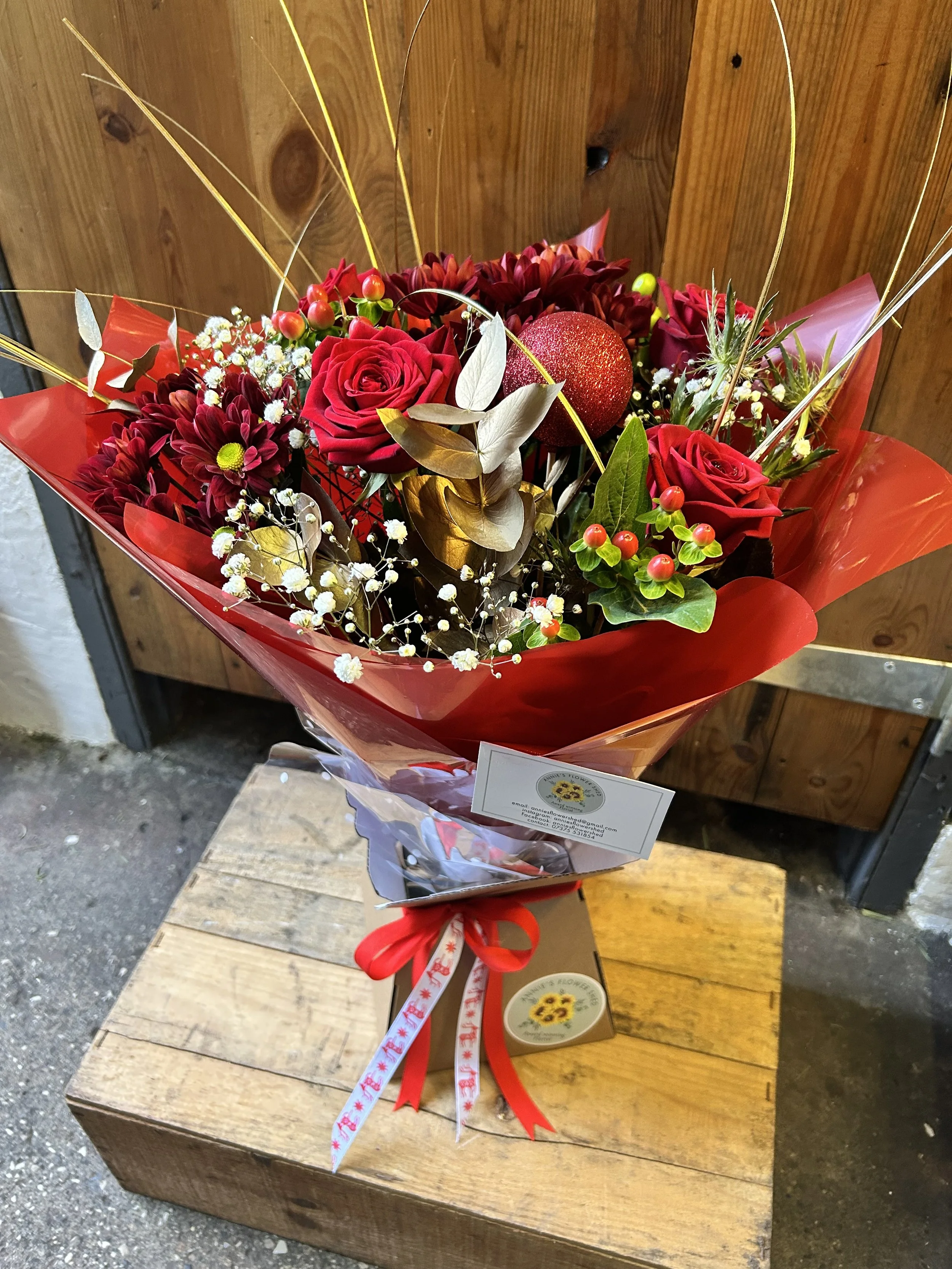 Bouquet of red roses, burgundy dahlias, small white and yellow flowers, green leaves, and decorative gold and white accents wrapped in red paper with ribbon ties, resting on a wooden surface.
