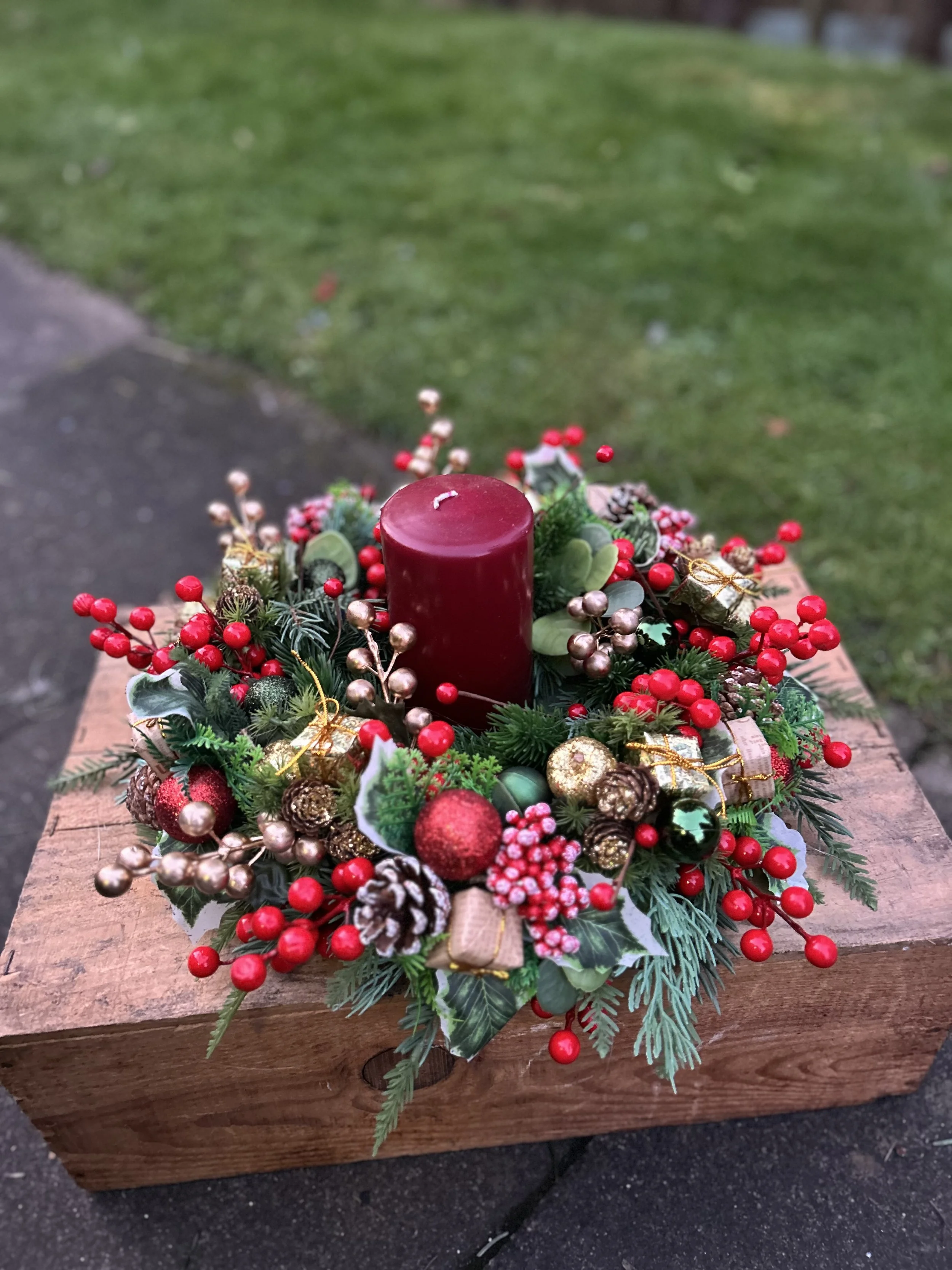 A holiday centerpiece with a red candle in the middle, surrounded by Christmas ornaments, greenery, and red berries, placed on a wooden surface outdoor.