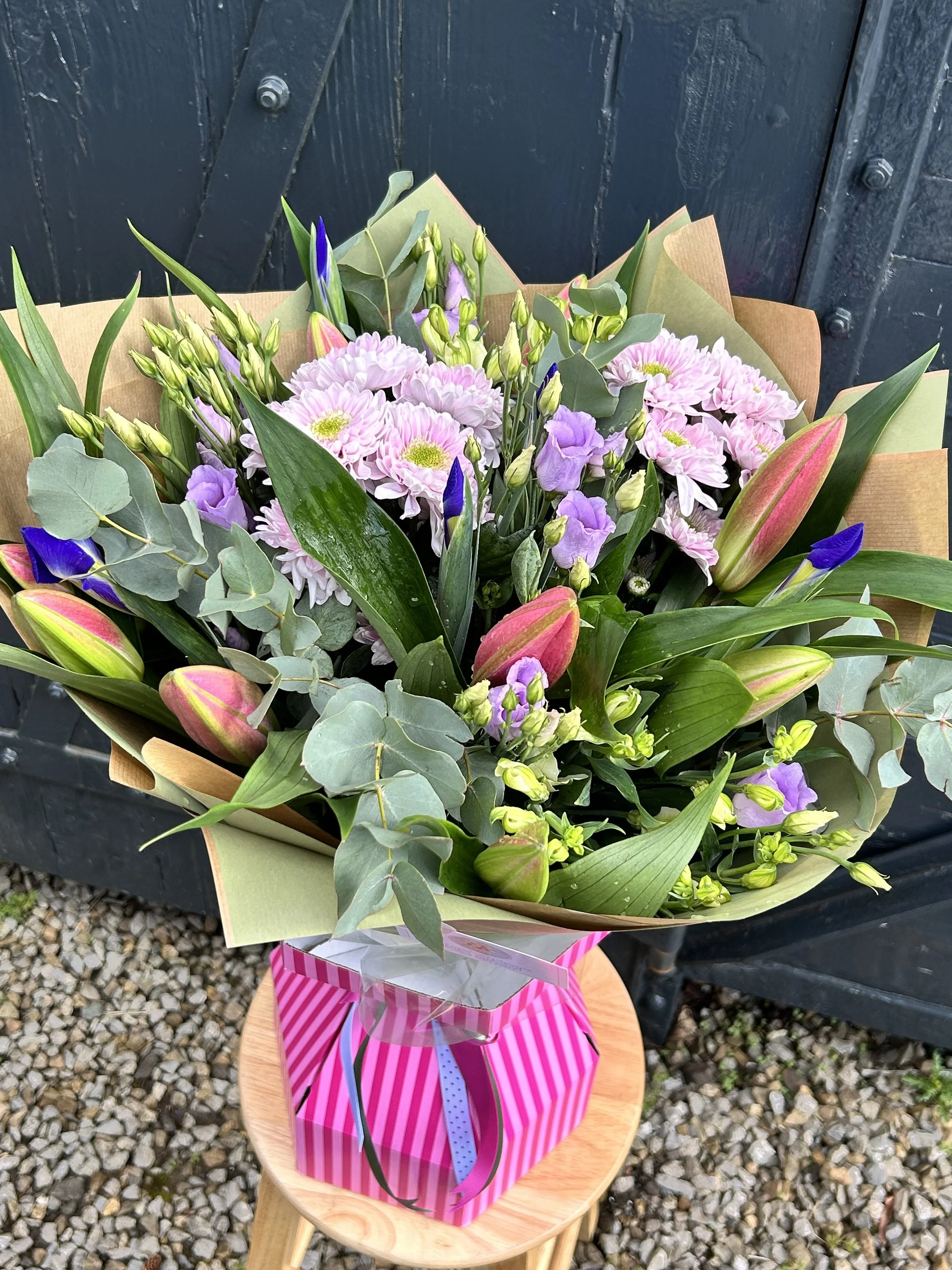A colorful bouquet of pink, purple, and green flowers wrapped in decorative paper and sitting on a wooden stool outdoors.