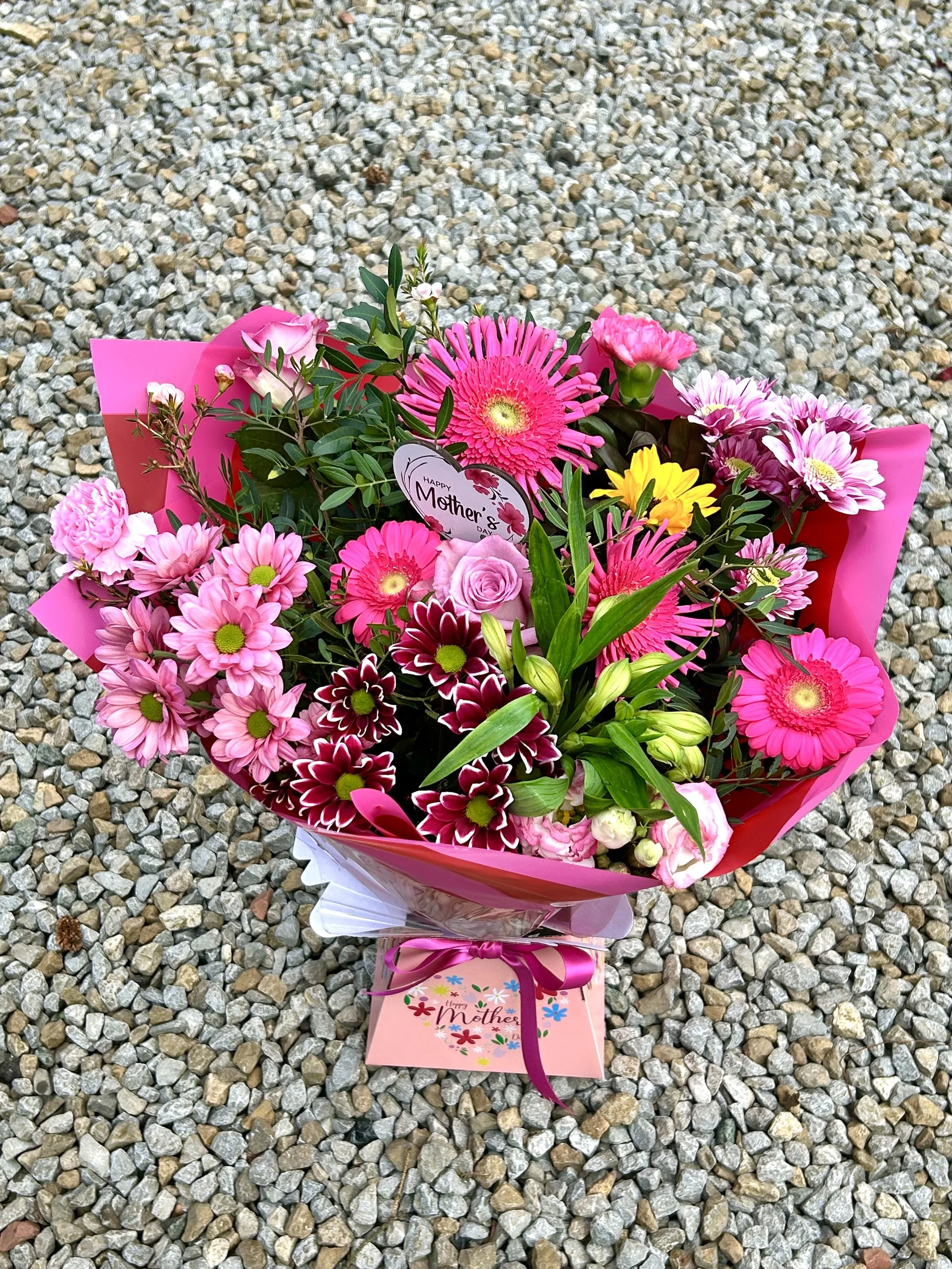 A colorful Mother's Day flower bouquet on a gravel ground with a pink tag that says 'Happy Mother' and a card with floral designs and a pink ribbon.