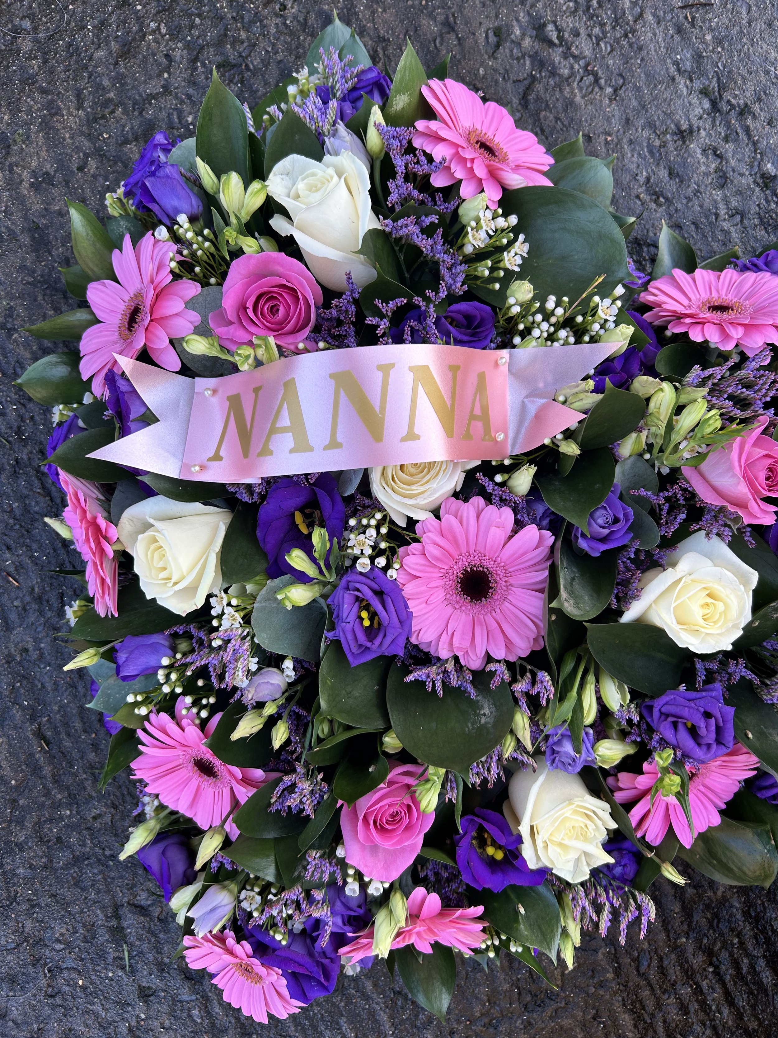 Colorful floral arrangement with pink, white, purple, and lavender flowers on a dark asphalt background, featuring a pink ribbon with the word 'NANA' in gold.