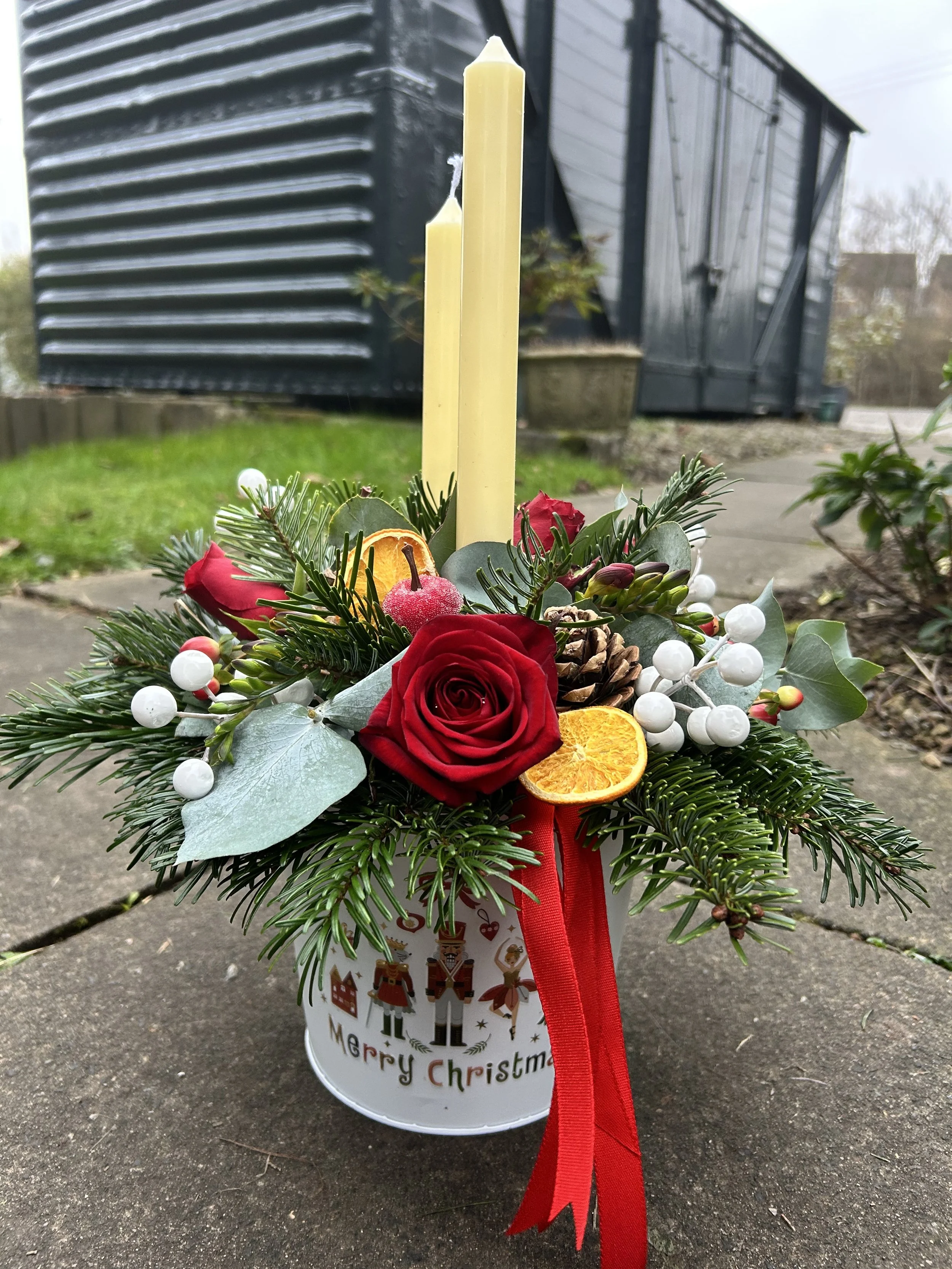 A festive Christmas arrangement with red roses, evergreen branches, dried orange slices, white berries, a pine cone, and a red ribbon, placed in a white container with holiday-themed illustrations and the words 'Merry Christmas.'
