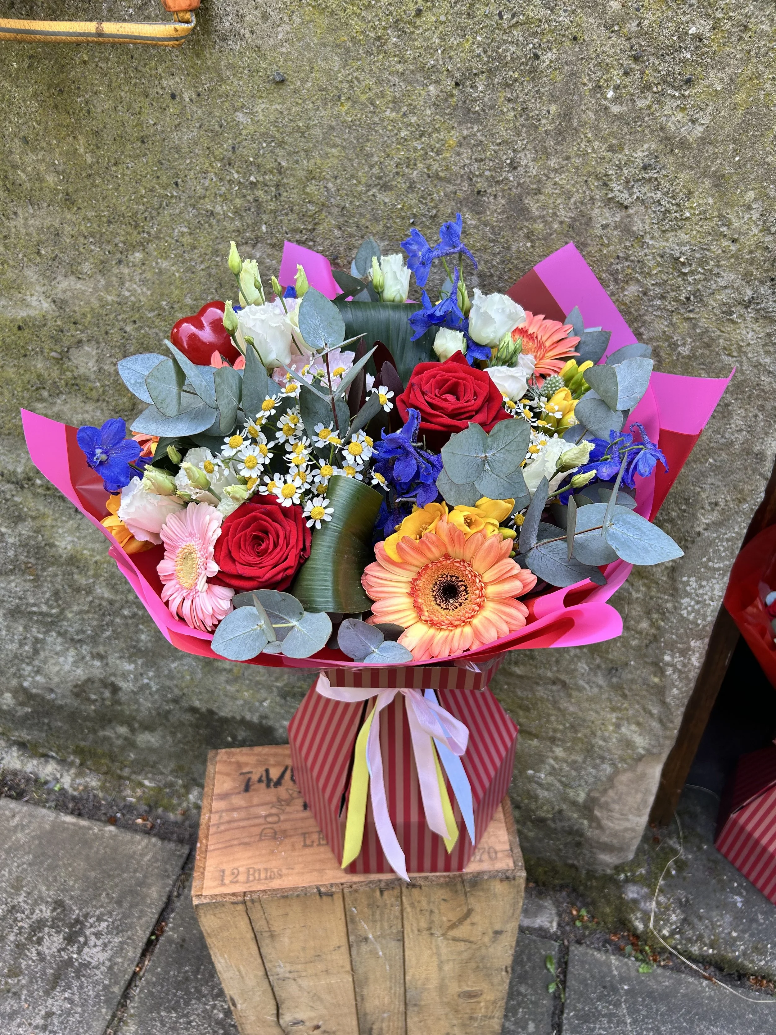 A colorful bouquet of flowers, including roses, daisies, and other blossoms, wrapped in pink paper with a striped ribbon, resting on a wooden crate outdoors against a mossy concrete wall.