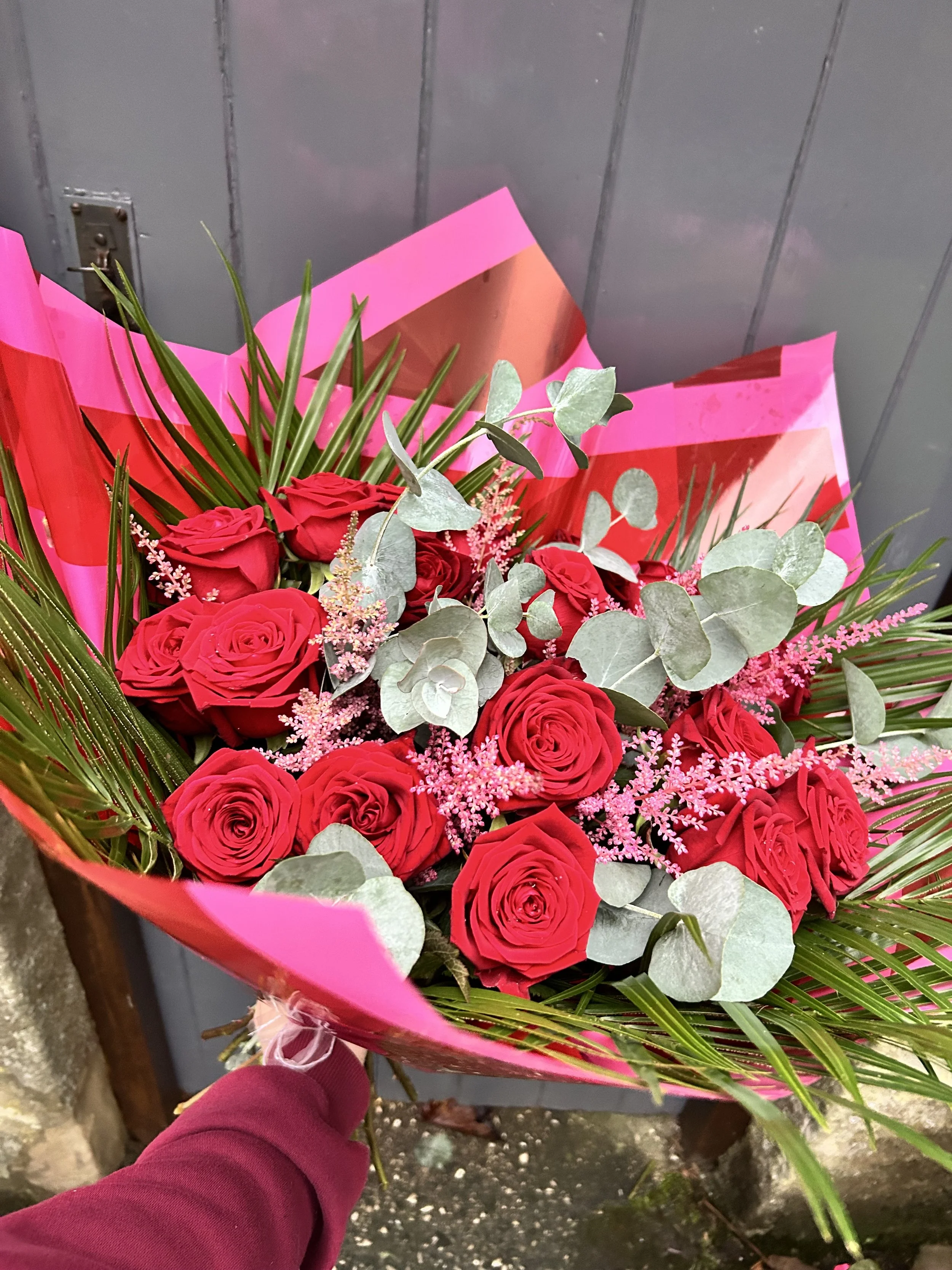 A bouquet of red roses, pink filler flowers, green leaves, and eucalyptus leaves wrapped in pink paper, being held by a person wearing a maroon sleeve.