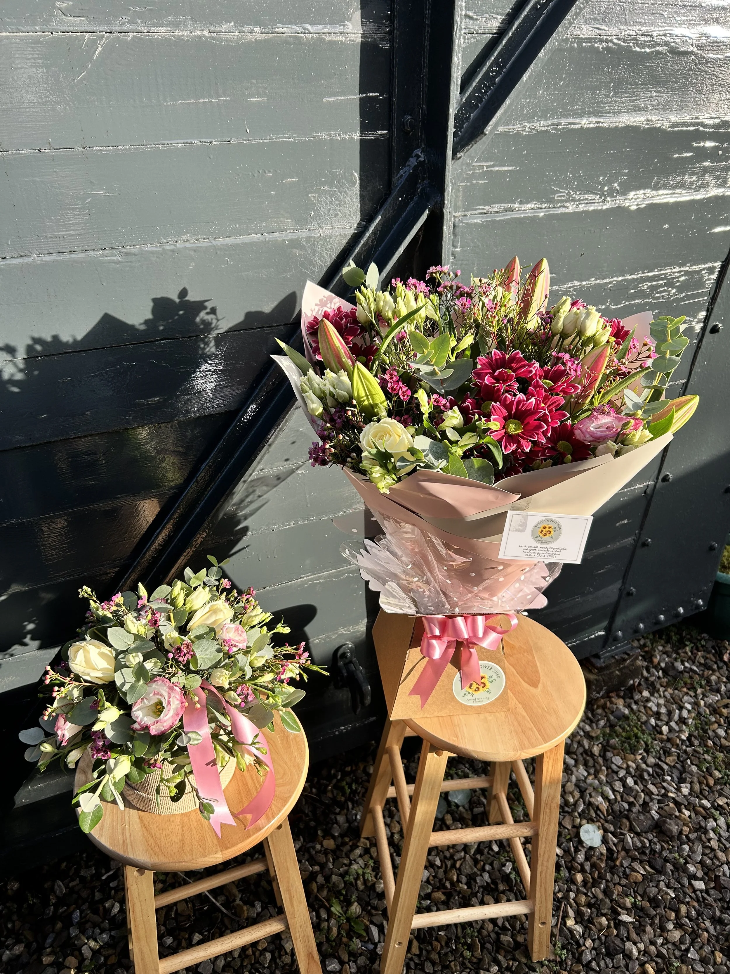 Two small wooden stools each with a floral arrangement on top, placed against a dark gray wooden wall; the arrangements include pink, white, and green flowers and are decorated with pink ribbons.