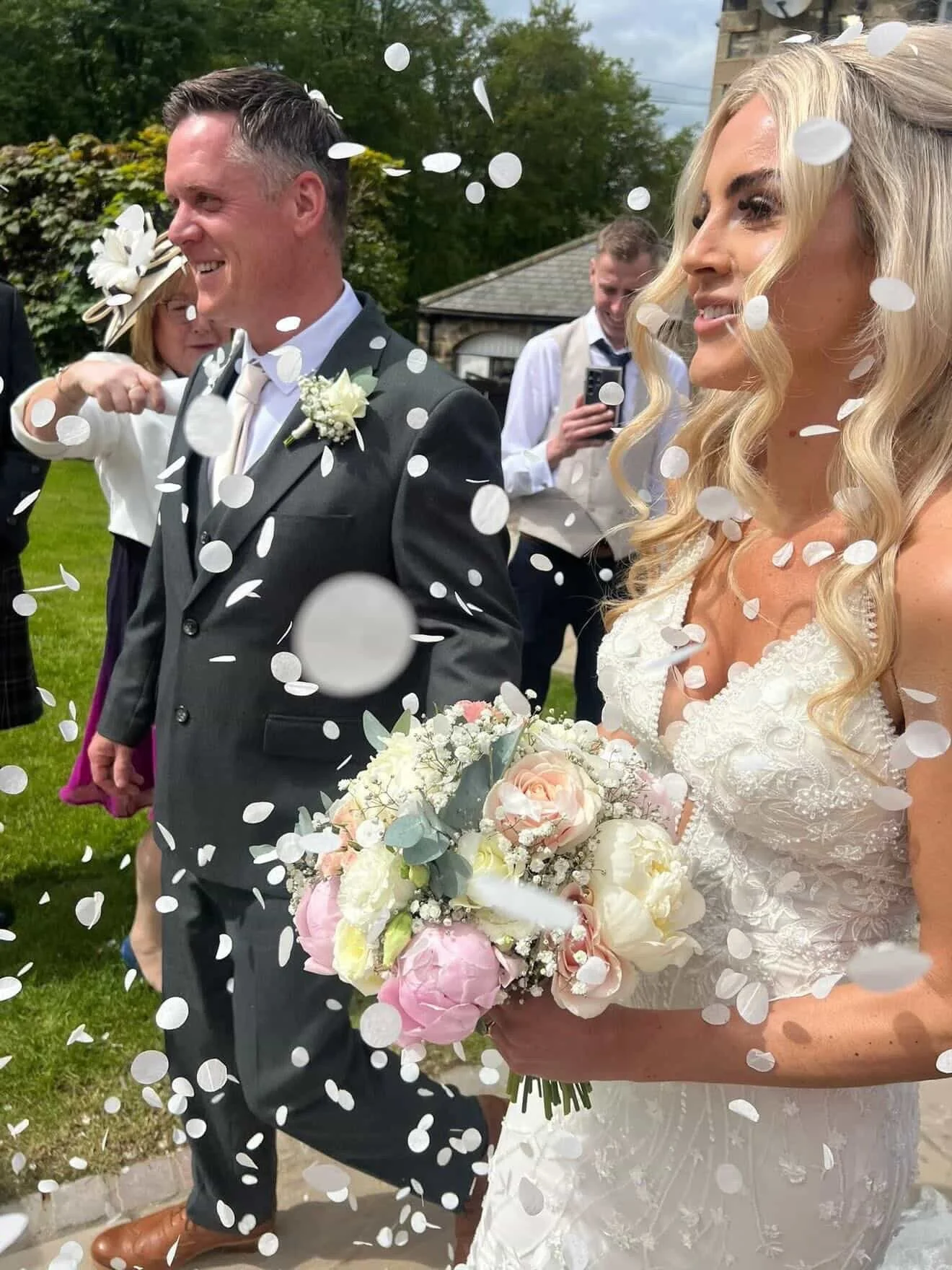 Bride holding a bouquet of pink and white flowers, standing next to groom, wedding guests throwing white confetti, outdoor wedding setting.
