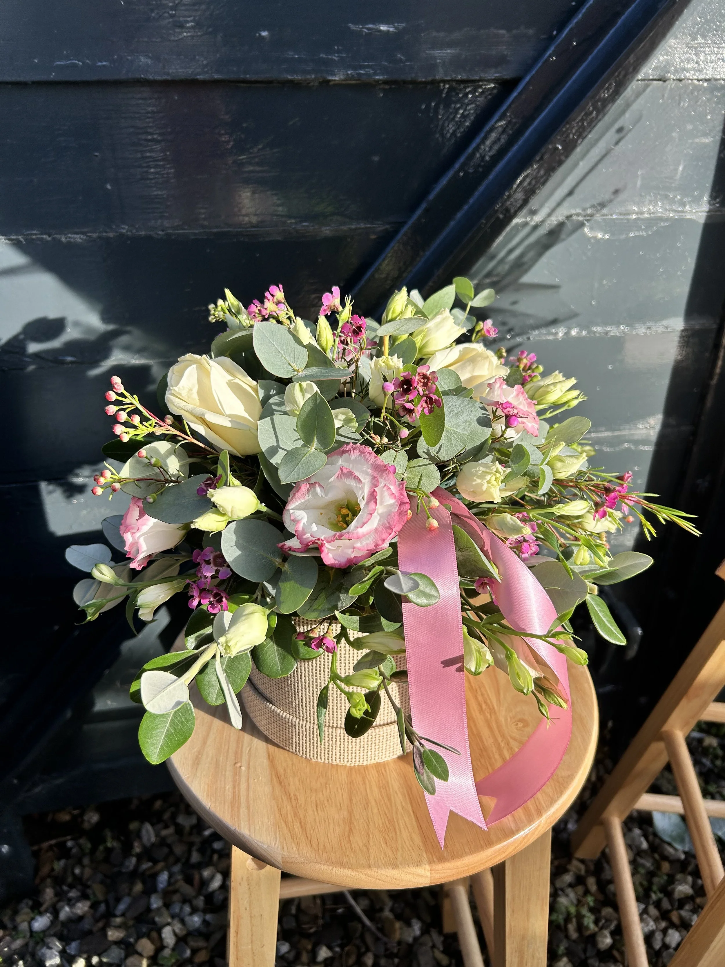 A bouquet of white and pink flowers with green leaves and pink ribbon in a beige basket on a round wooden table.