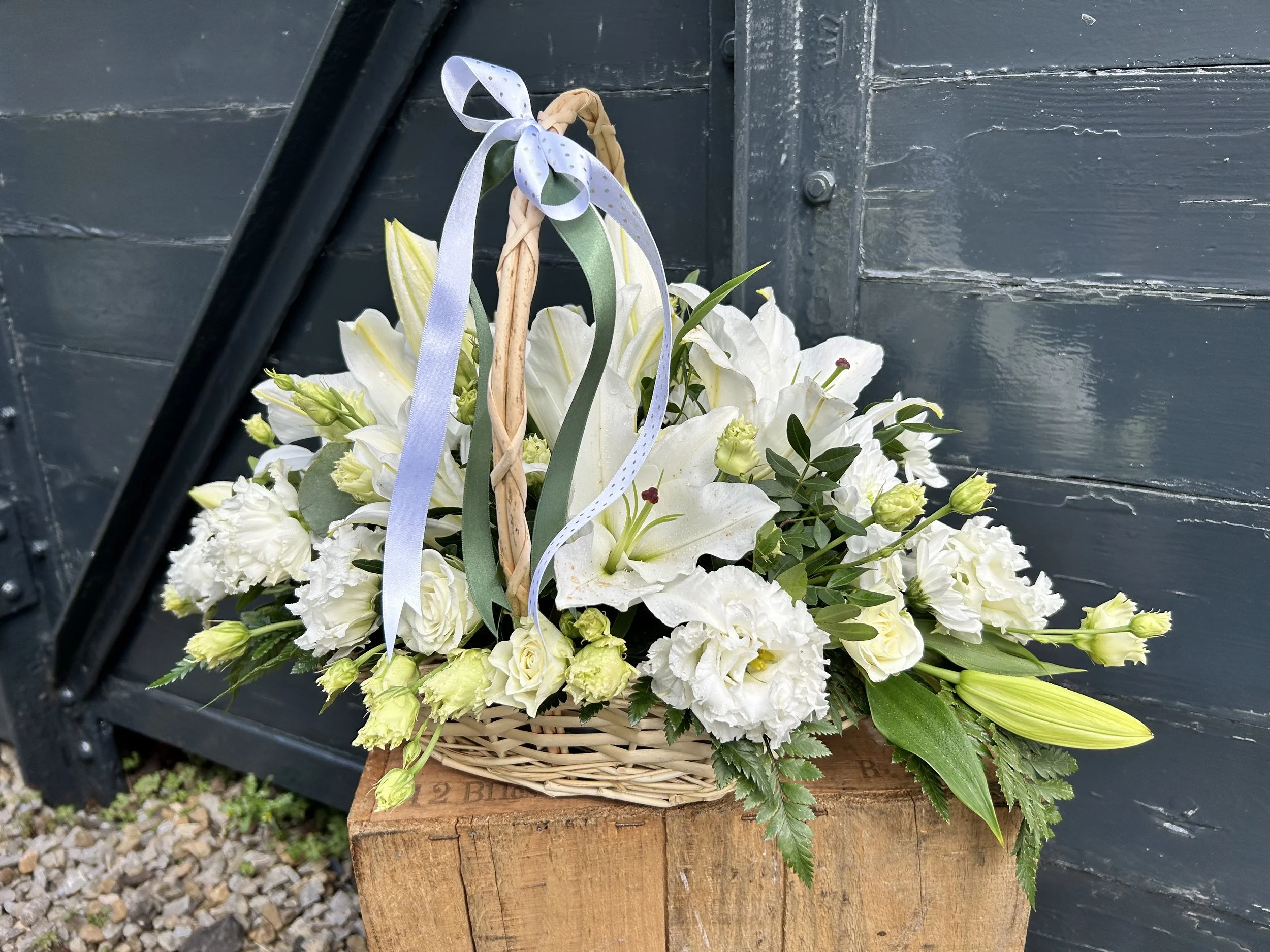 A basket of white lilies and other flowers placed on a wooden block against a dark wall with visible texture.