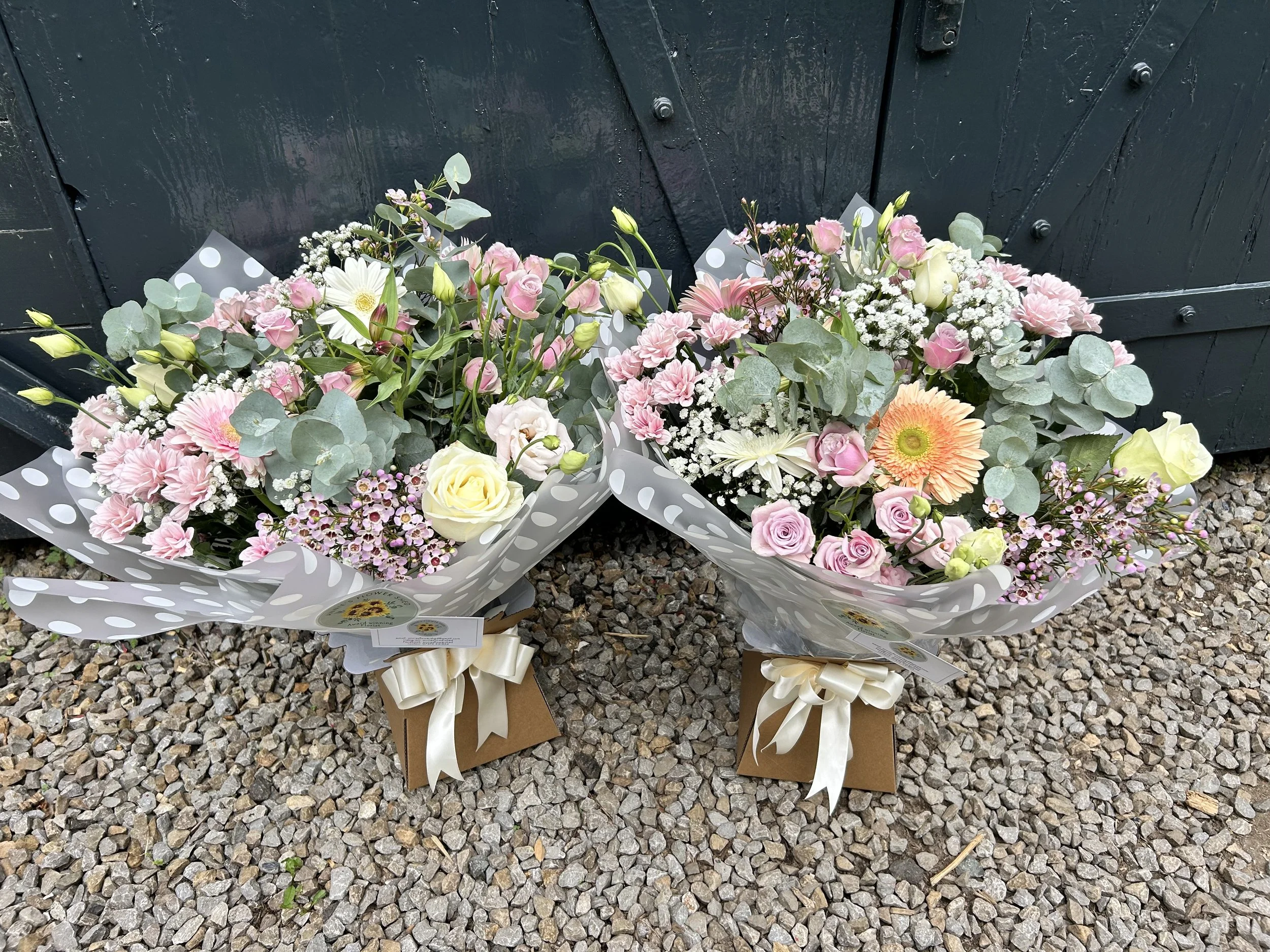 Two bouquets of pink and white flowers wrapped in polka dot paper, placed on a gravel surface in front of a black wooden fence.