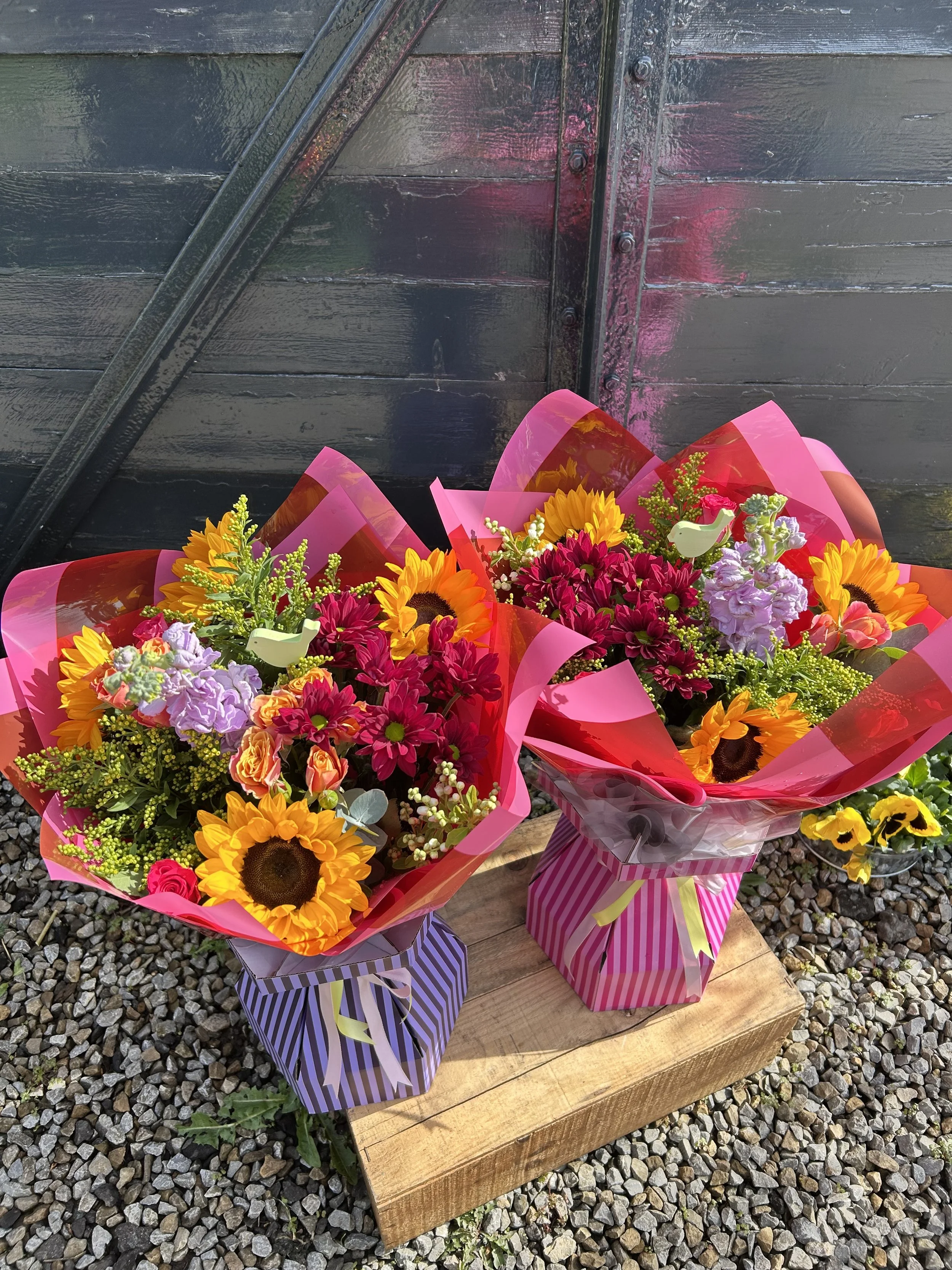 Three colorful flower bouquets wrapped in pink, purple, and clear cellophane are placed on a wooden surface outdoors against a dark wooden wall with metal details and pink paint streaks.