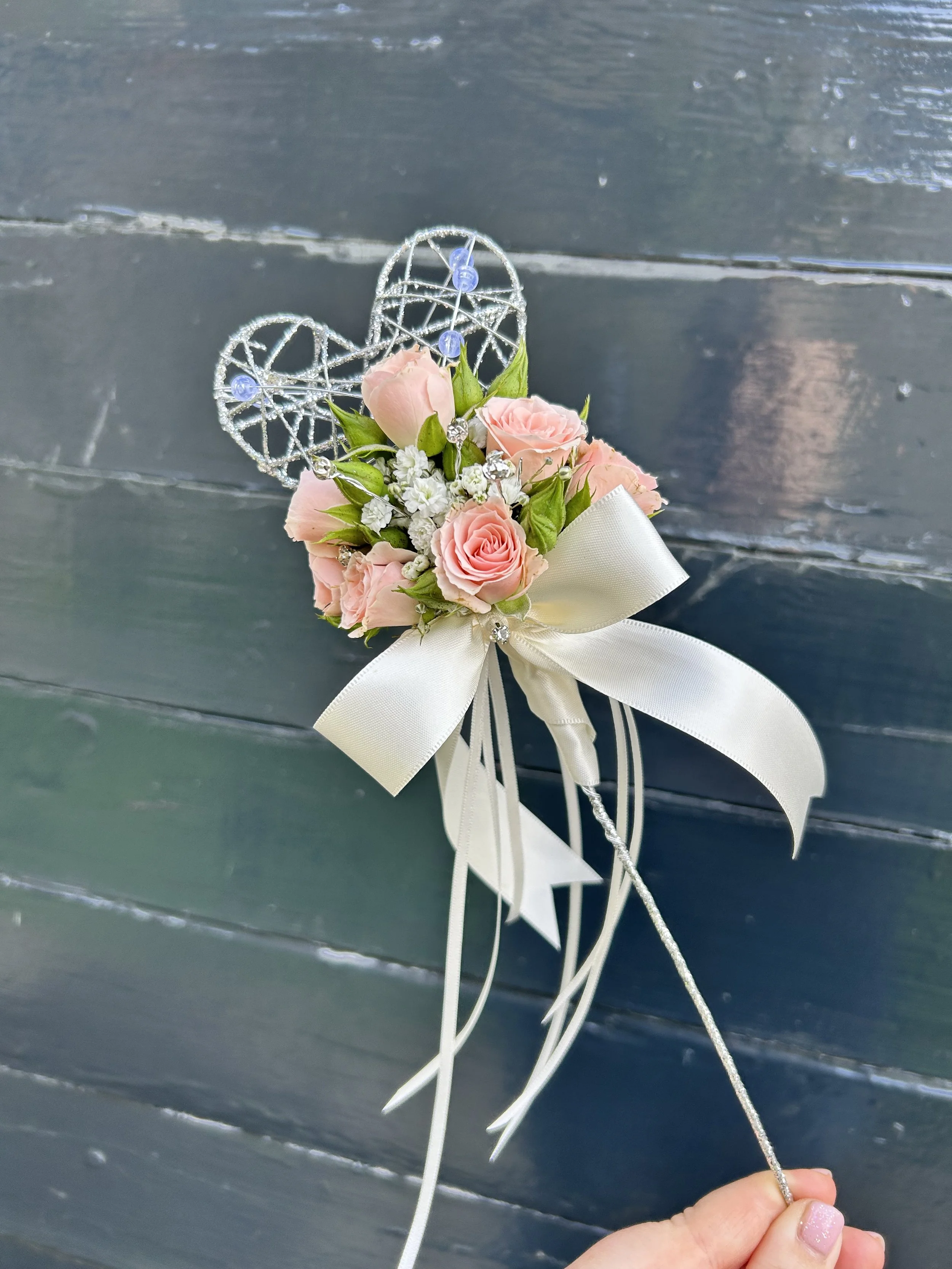 A hand holding a decorative stick with pink roses, white baby's breath flowers, green leaves, and a white ribbon, with a blue and silver wire heart ornament attached.