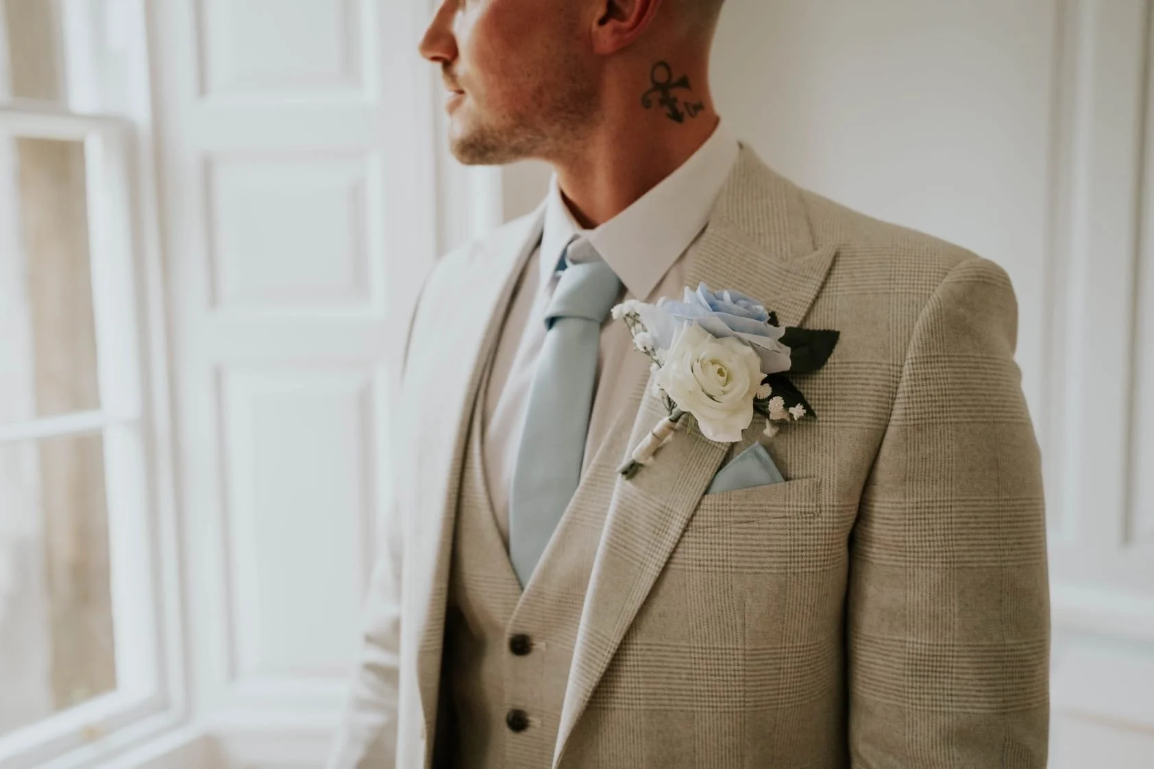 Close-up of a man in a beige checked suit with a boutonnière, standing indoors near a window.