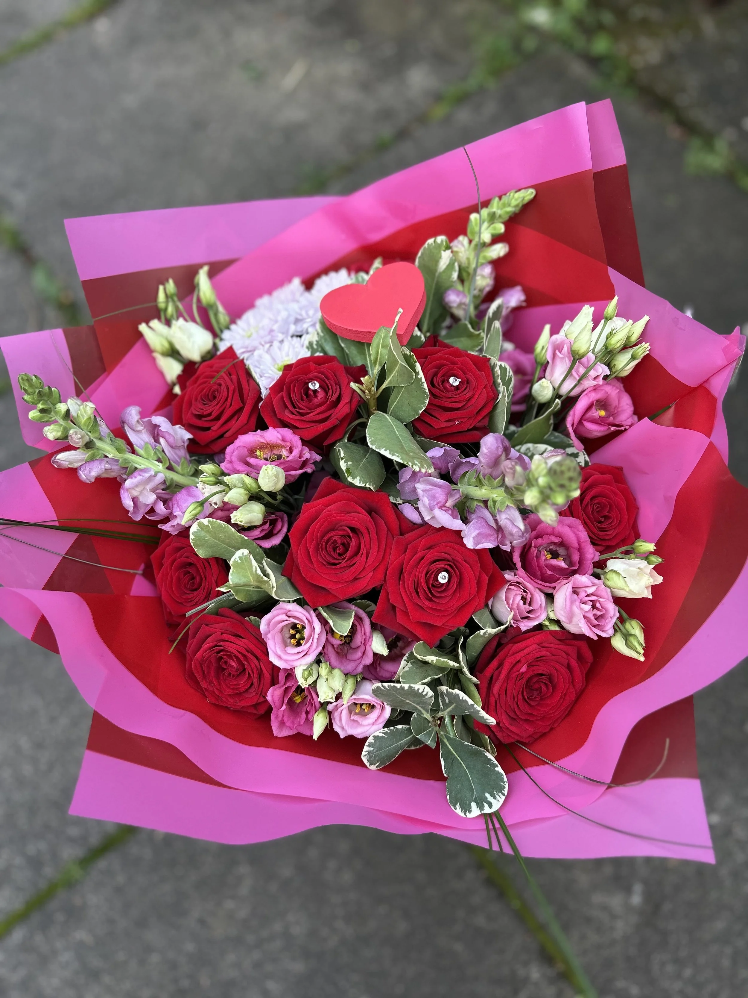 Bouquet of red roses, pink flowers, and greenery wrapped in pink and red paper, with a small red heart decoration, on a concrete surface.