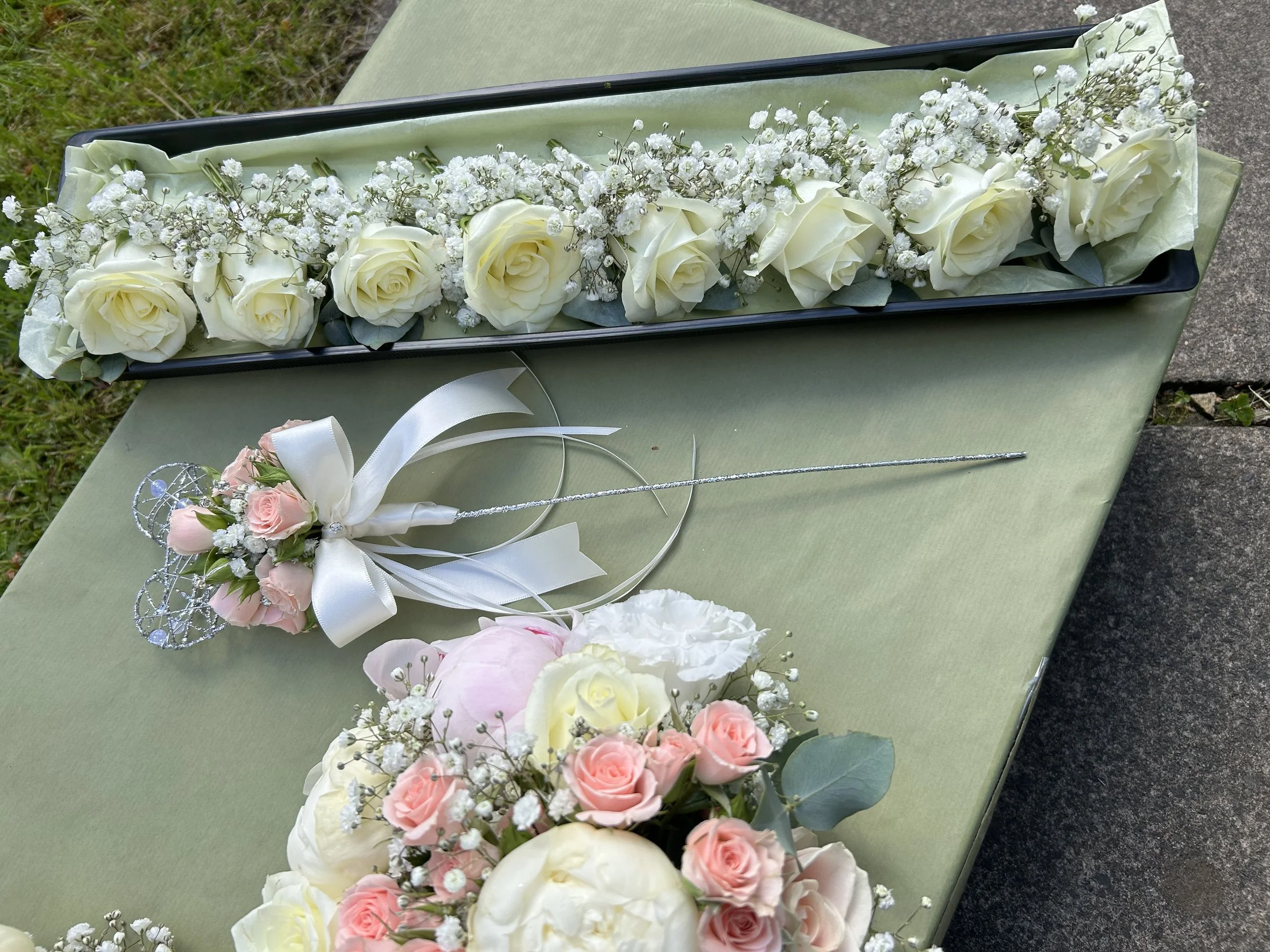 Arrangement of white roses and baby's breath flowers in a black box on a light green table outdoors.