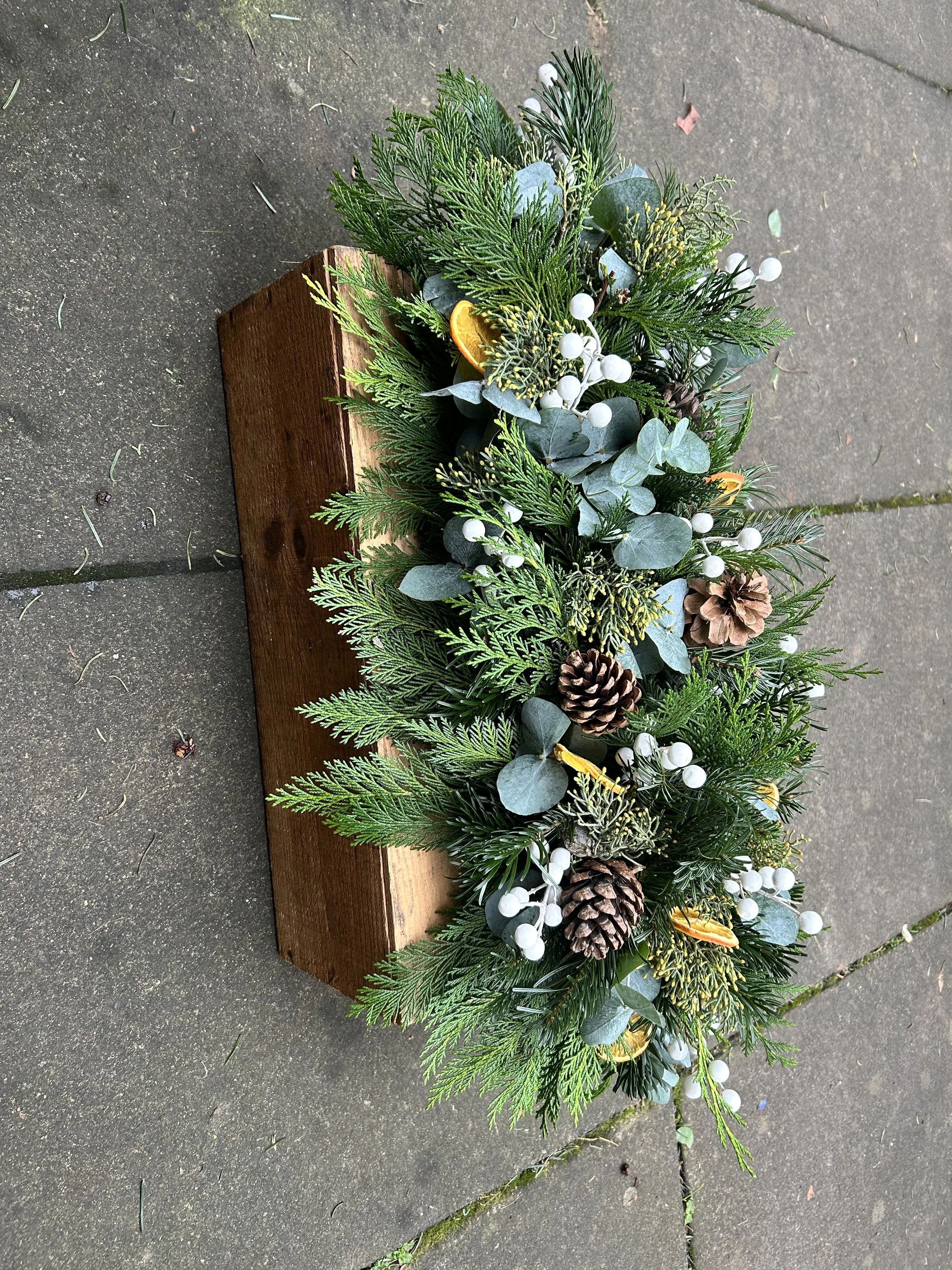 A wooden table decorated with a holiday arrangement of green pine, eucalyptus, white berries, and pinecones on a concrete surface.