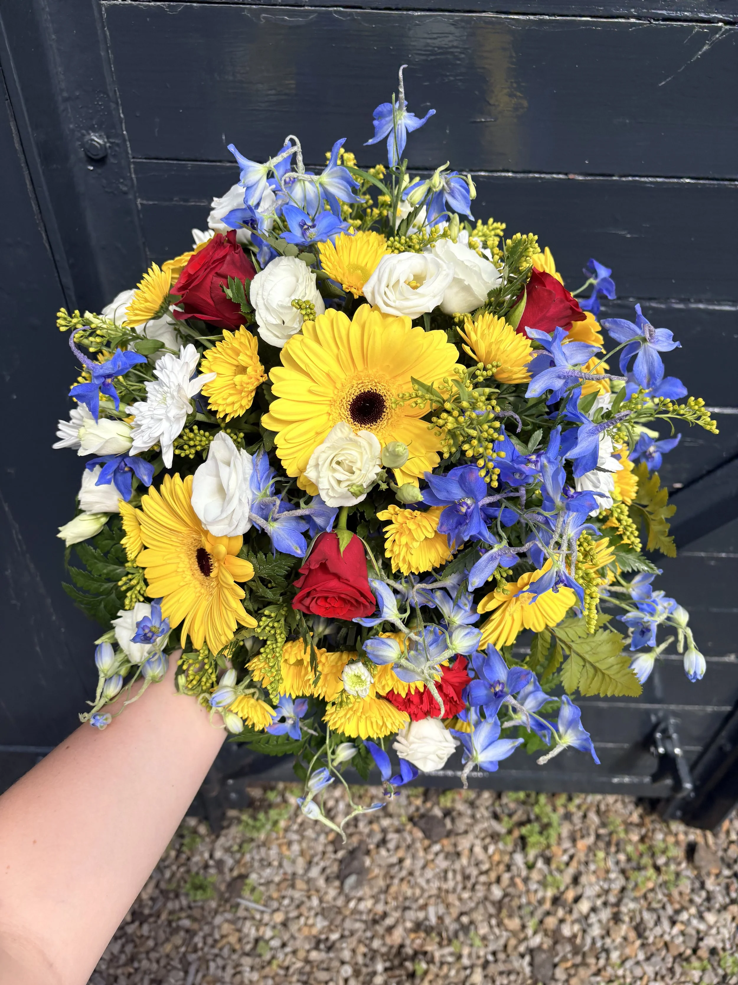 A colorful bouquet of various flowers including yellow daisies, white roses, red roses, and blue delphiniums, held by a person's hand against a dark outdoor background.
