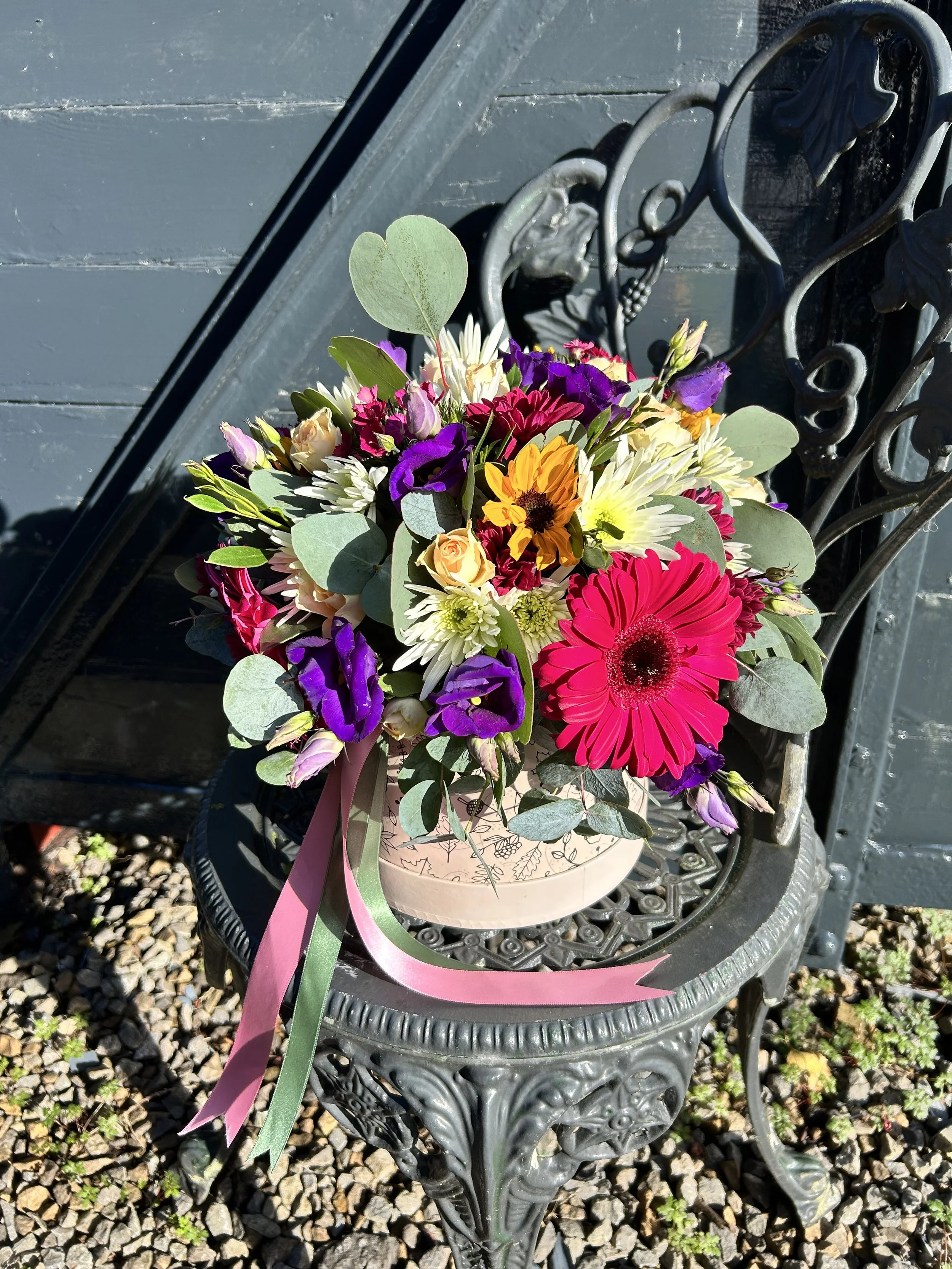 A colorful bouquet of flowers, including pink, purple, yellow, and white blooms, arranged in a light pink gift box with ribbons, placed on a decorative metal outdoor table with a curved design, against a background of gravel and a gray wall.
