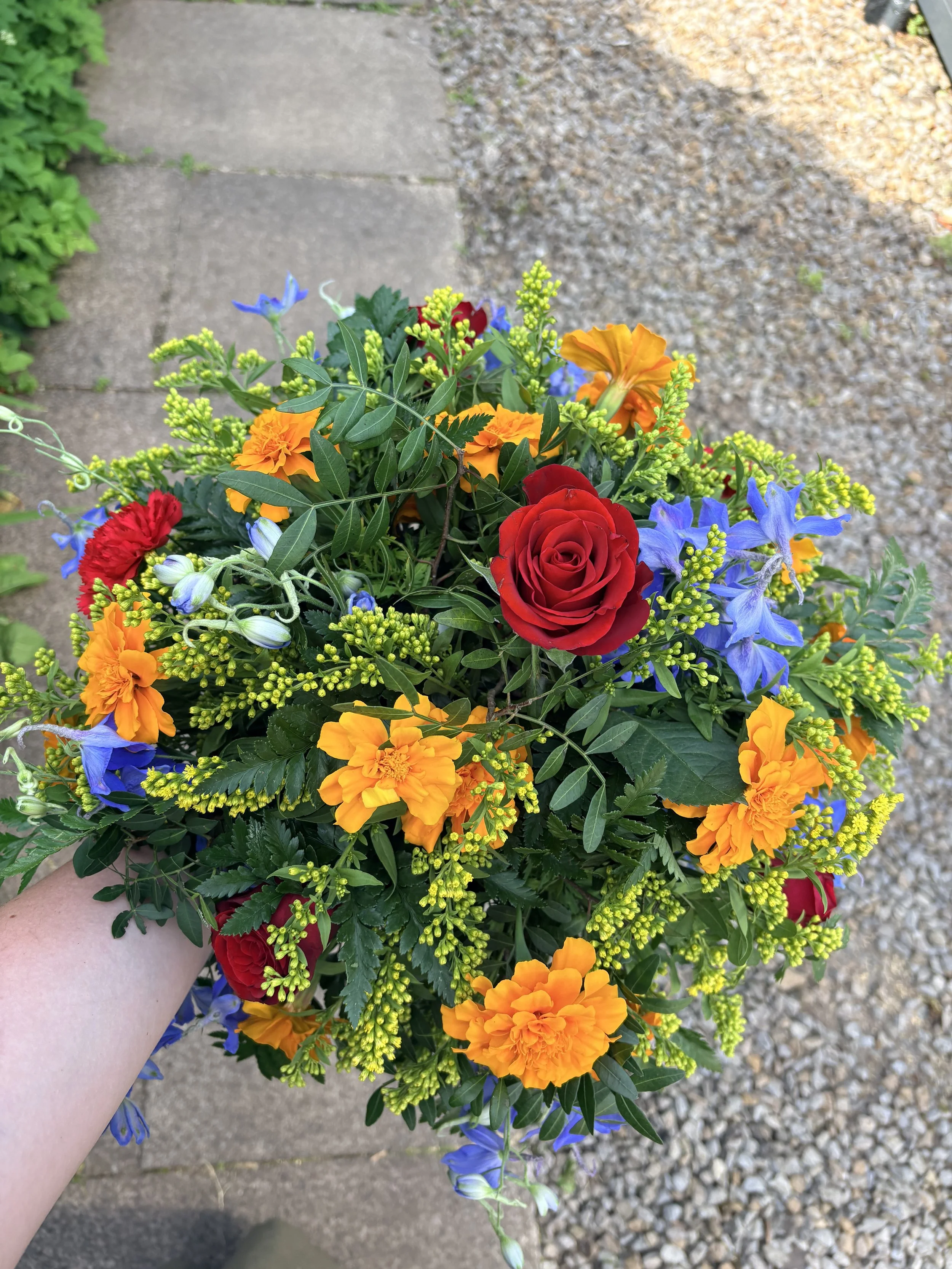 A diverse bouquet of flowers including red roses, orange marigolds, blue delphiniums, small yellow flowers, and various green foliage, being held outdoors over a pathway with stone and gravel.