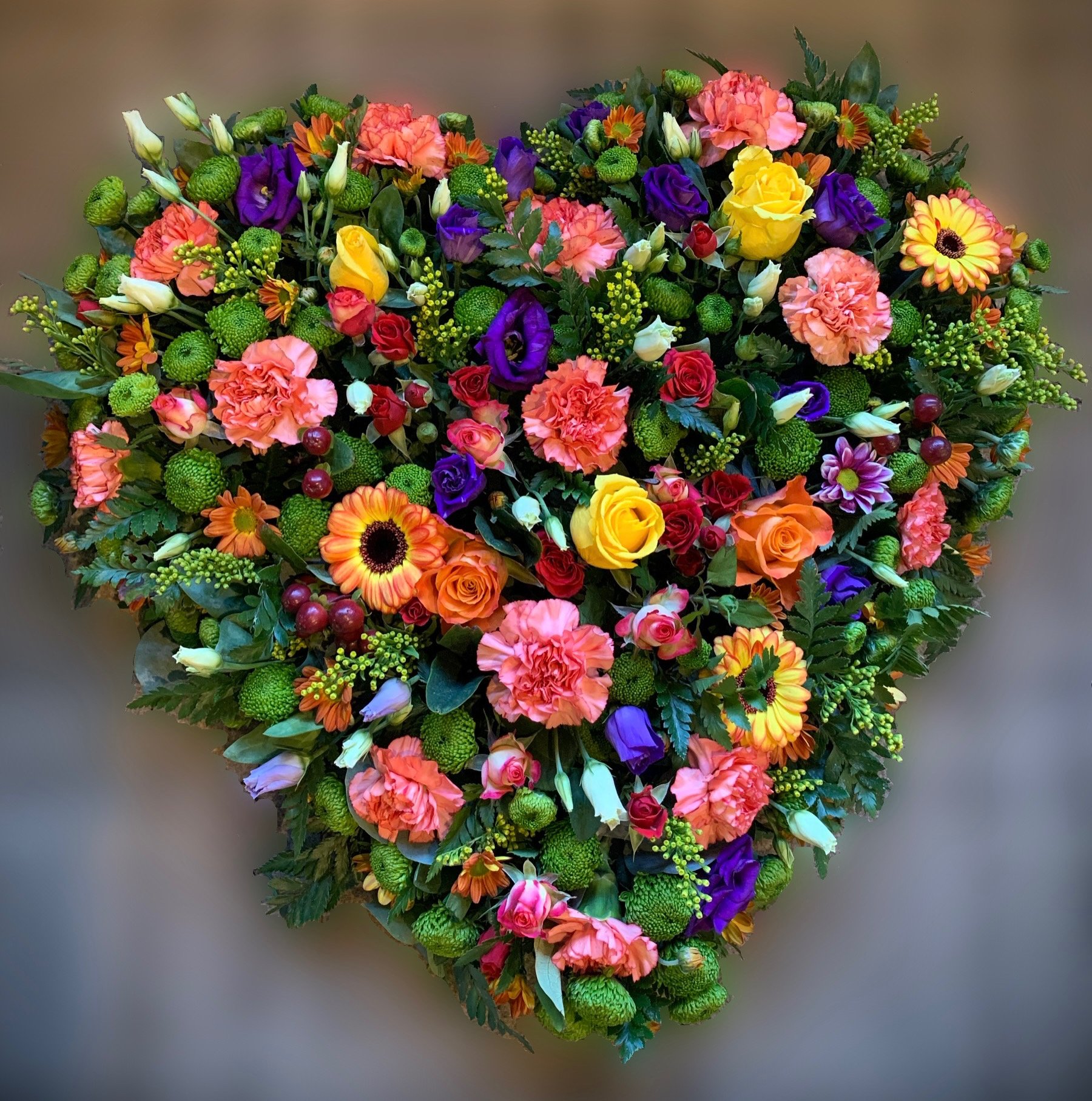 Heart-shaped floral arrangement with pink, yellow, orange, purple, white, and red flowers, including roses, carnations, gerbera daisies, lisianthus, and greenery.
