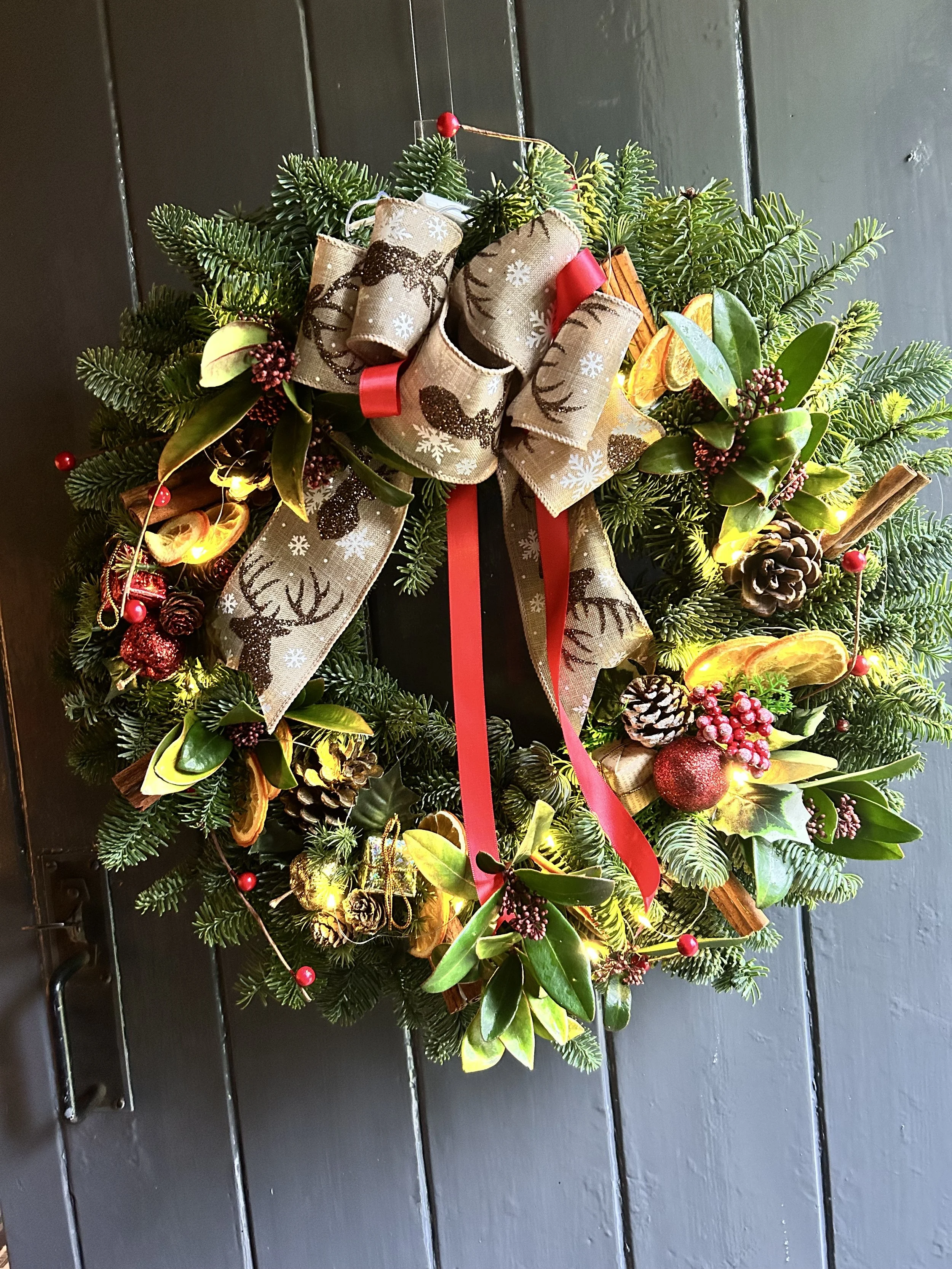A Christmas wreath decorated with sprigs of greenery, pinecones, dried orange slices, berries, a beige bow with reindeer and snowflakes, and red ribbons hanging on a dark door.