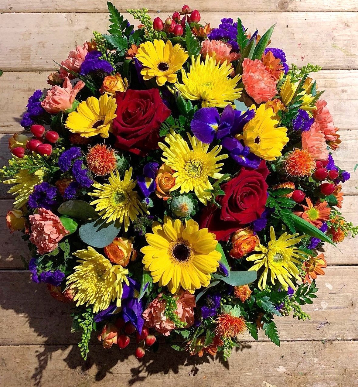 A colorful bouquet of various flowers including red roses, yellow gerbera daisies, purple irises, orange carnations, and berries, arranged on a wooden surface.