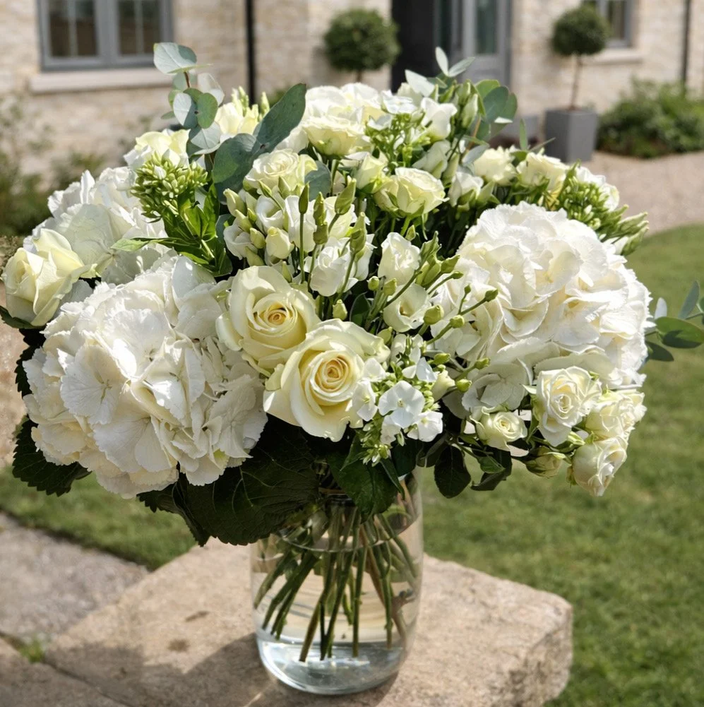 A bouquet of white flowers, including roses and hydrangeas, in a glass vase outdoors on a stone surface with a house and garden in the background.
