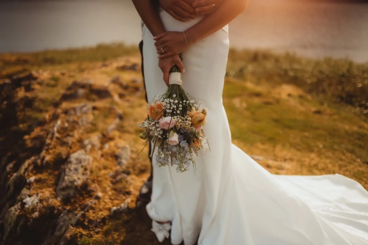 Person in white wedding dress holding a bouquet of flowers on a rocky outdoor landscape.