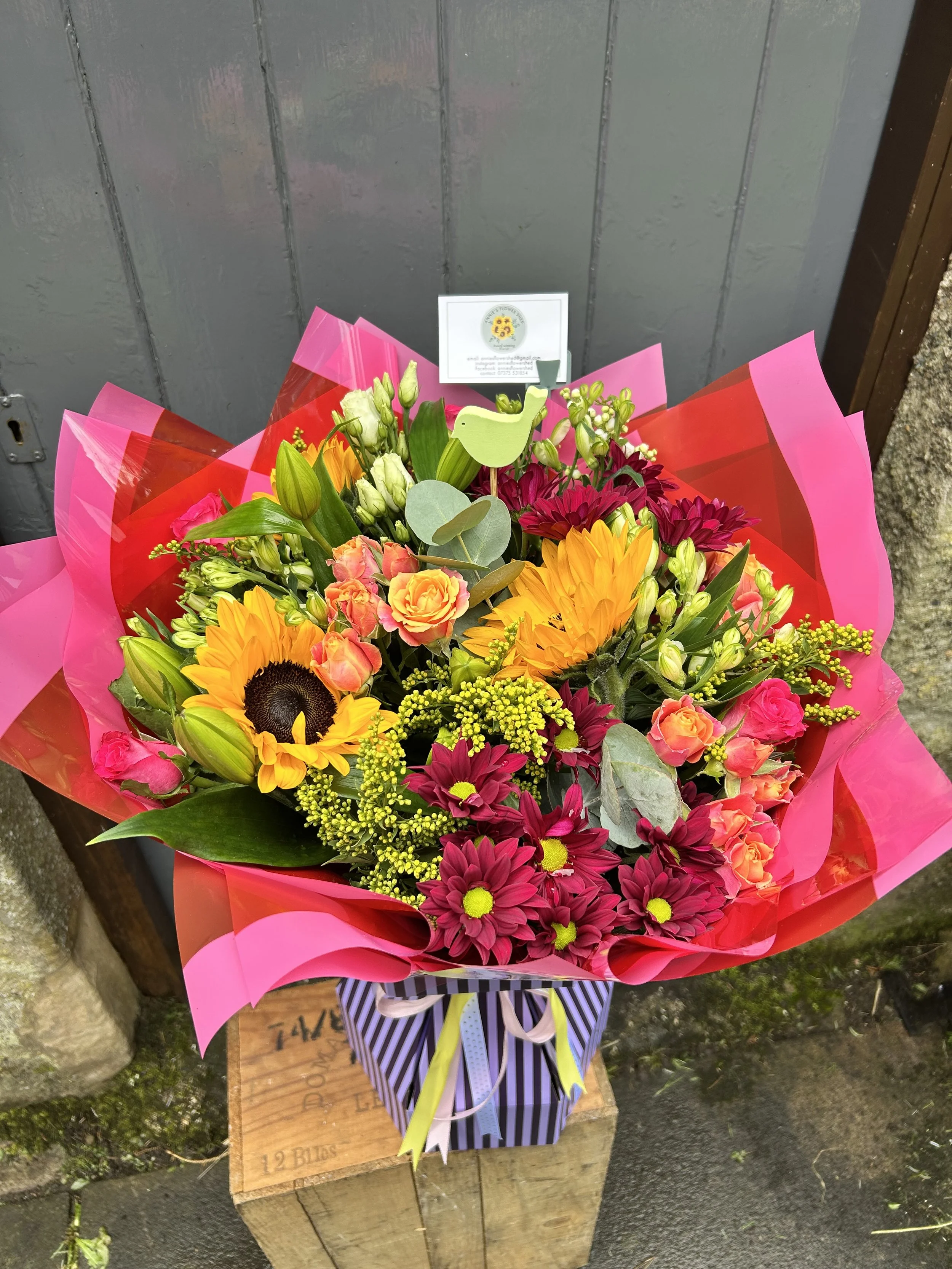 A colorful bouquet of flowers, including sunflowers, roses, and daisies, wrapped in pink and red paper, with a decorative ribbon, placed on a wooden stand outdoors.