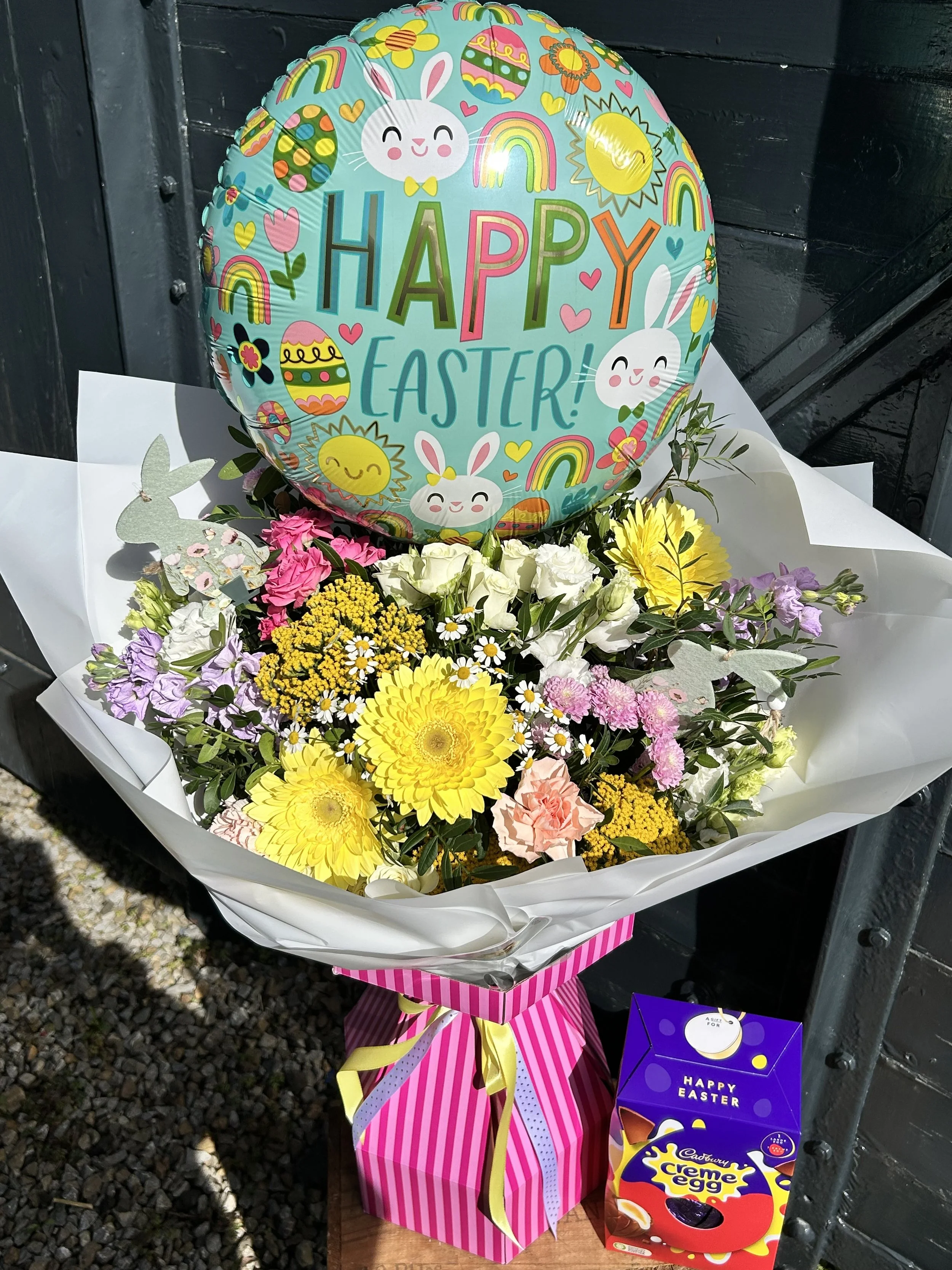 Easter-themed balloon with the words "HAPPY EASTER!" surrounded by colorful designs like bunnies, rainbows, sun, hearts, and decorated eggs, placed amidst a bouquet of yellow, pink, white, and purple flowers, with a purple Cadbury Creme Egg box nearb
