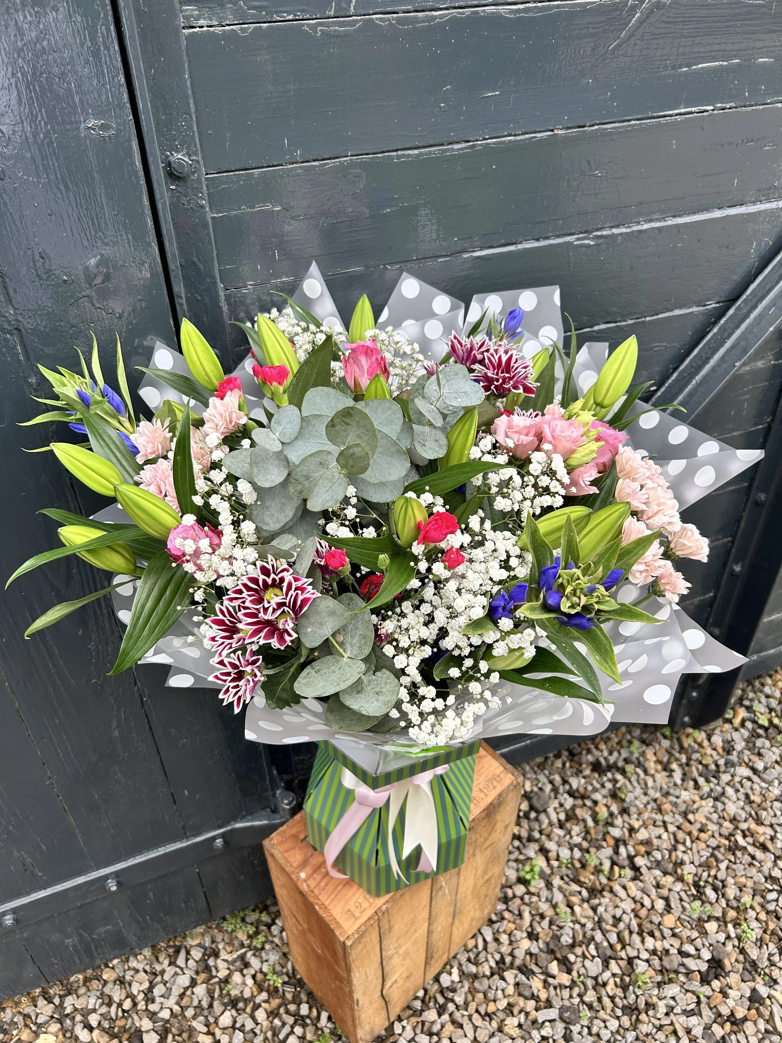A colorful bouquet of flowers wrapped in decorative paper and tied with a pink and green ribbon, resting on a wooden block on a gravel surface.
