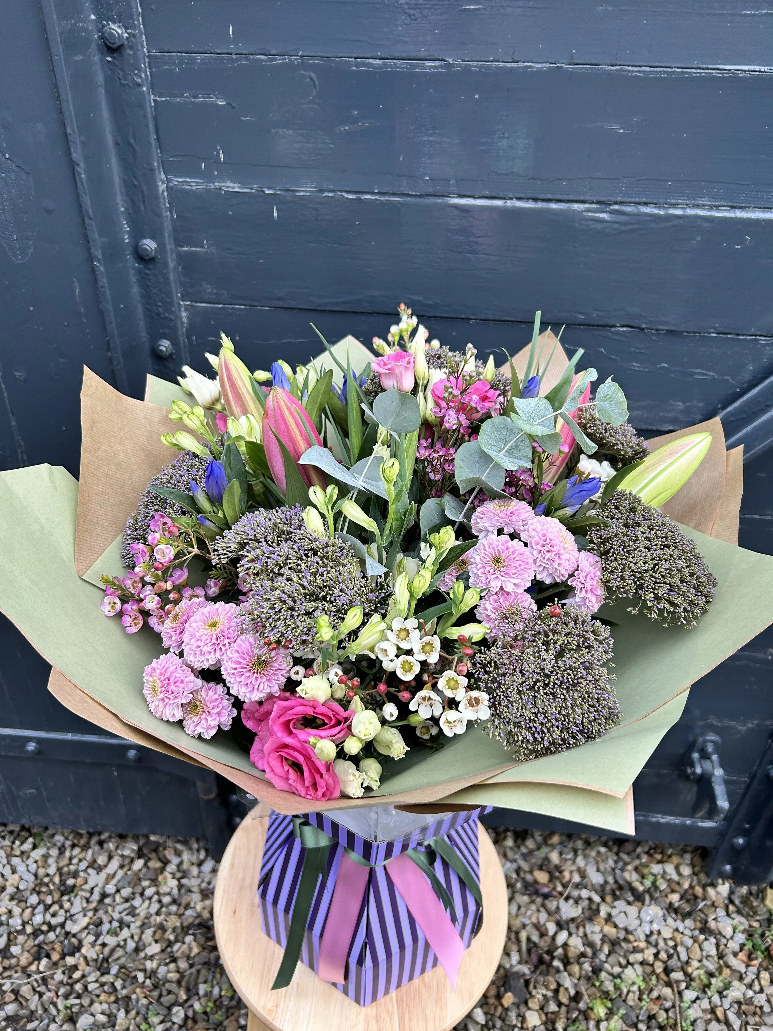 A bouquet of assorted pink, white, purple, and blue flowers wrapped in green and purple striped paper with pink, green, and purple ribbons, placed on a wooden table against a black background.