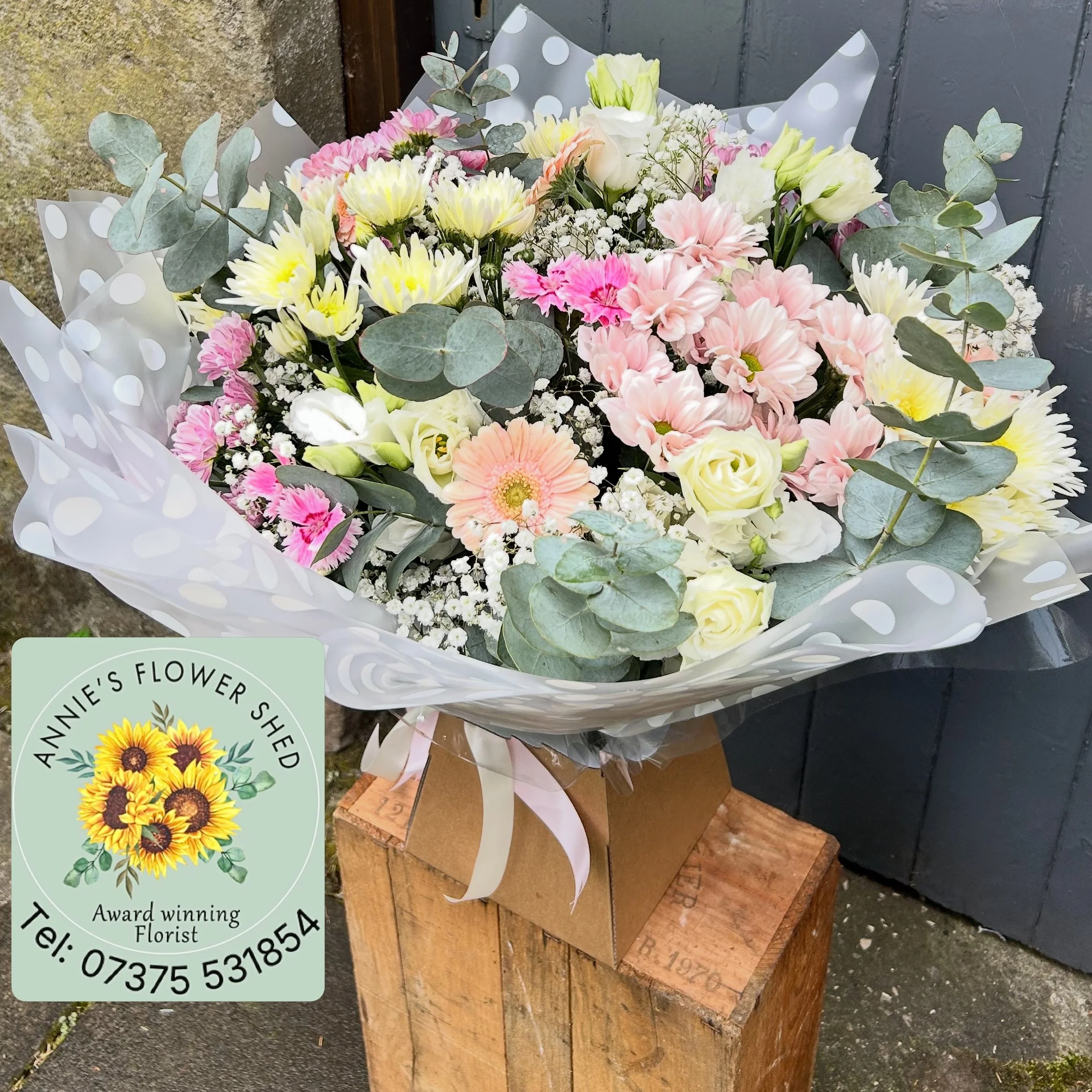 A bouquet of flowers includes white roses, pink carnations, light yellow daisies, baby's breath, and eucalyptus, wrapped in polka dot paper, resting on a wooden surface against a dark wall.