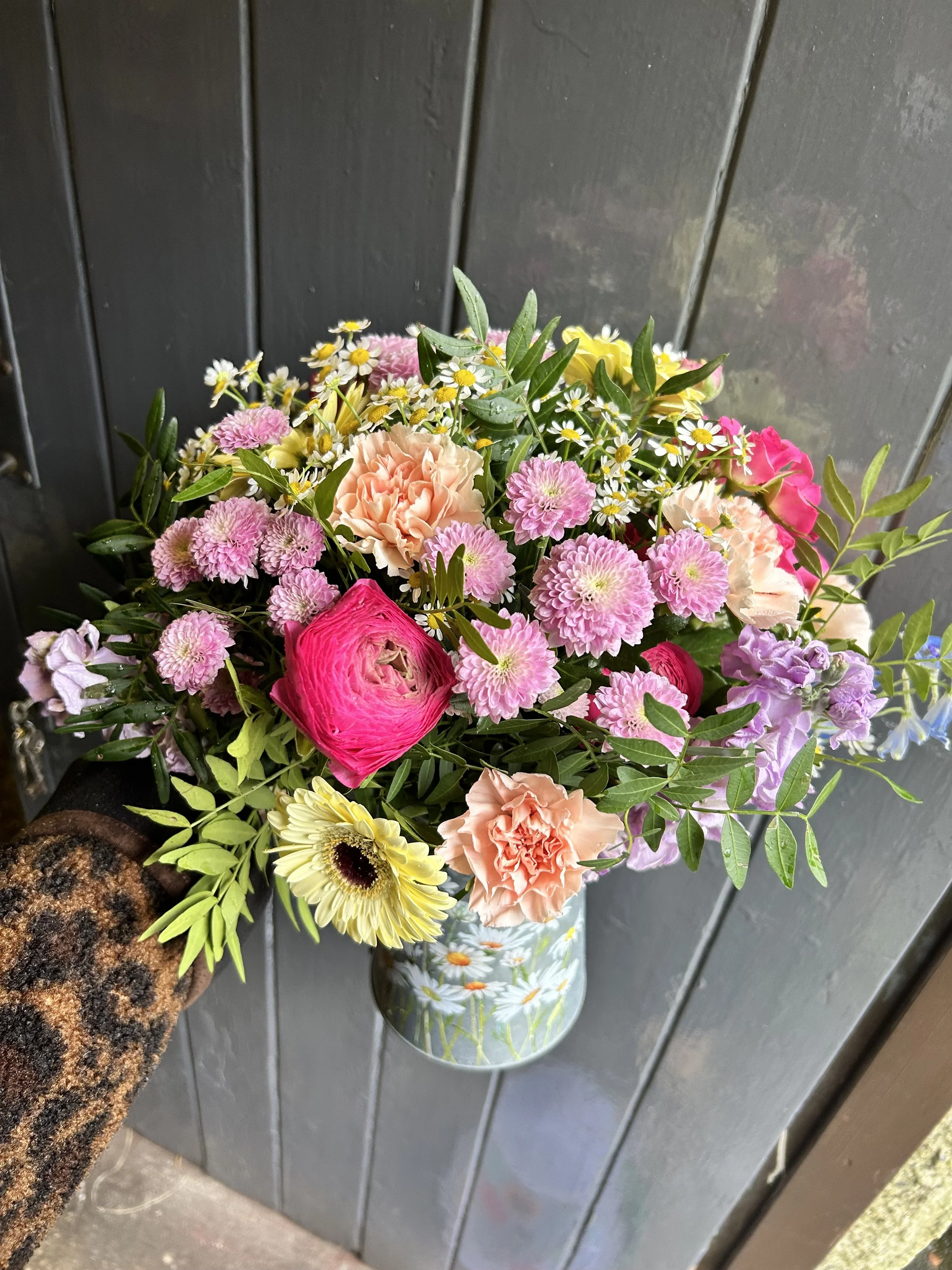 A colorful bouquet of flowers, including pink carnations, yellow daisies, and white daisies with yellow centers, in a glass vase on a dark wooden surface.
