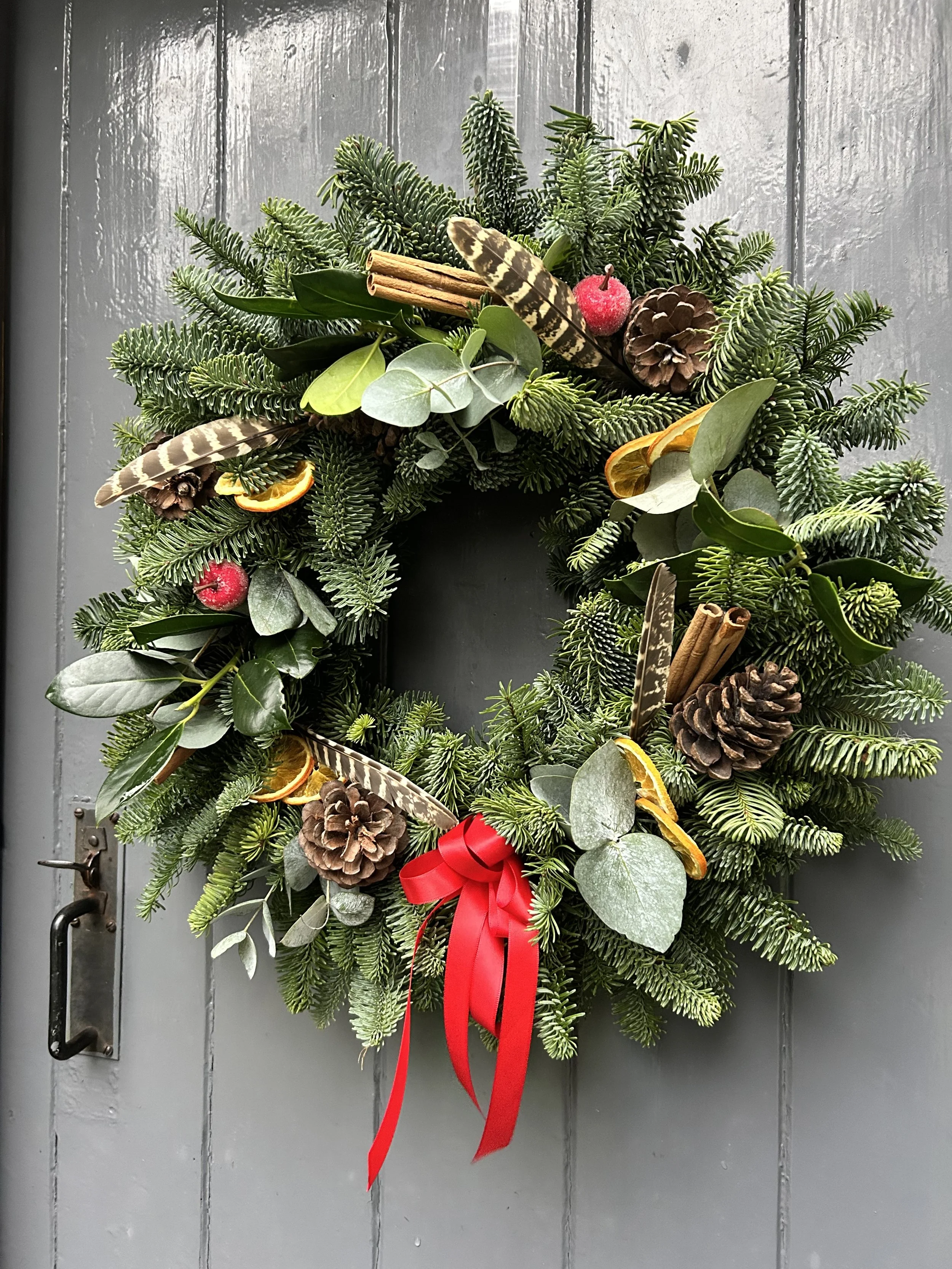 A Christmas wreath decorated with pine branches, pinecones, cinnamon sticks, dried orange slices, green leaves, a red berry, and a red bow, hanging on a gray door.