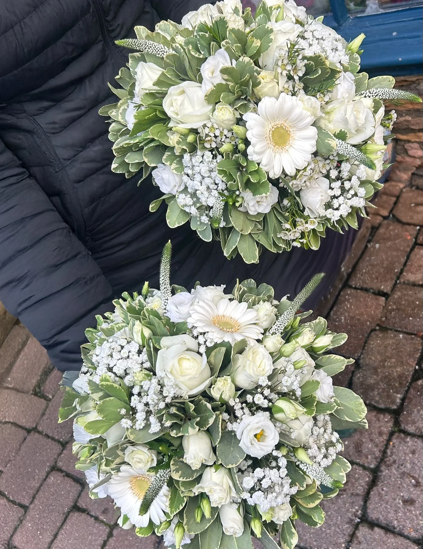 Two large bouquets of white flowers with green leaves, one resting on a black padded surface and the other on a brick surface.