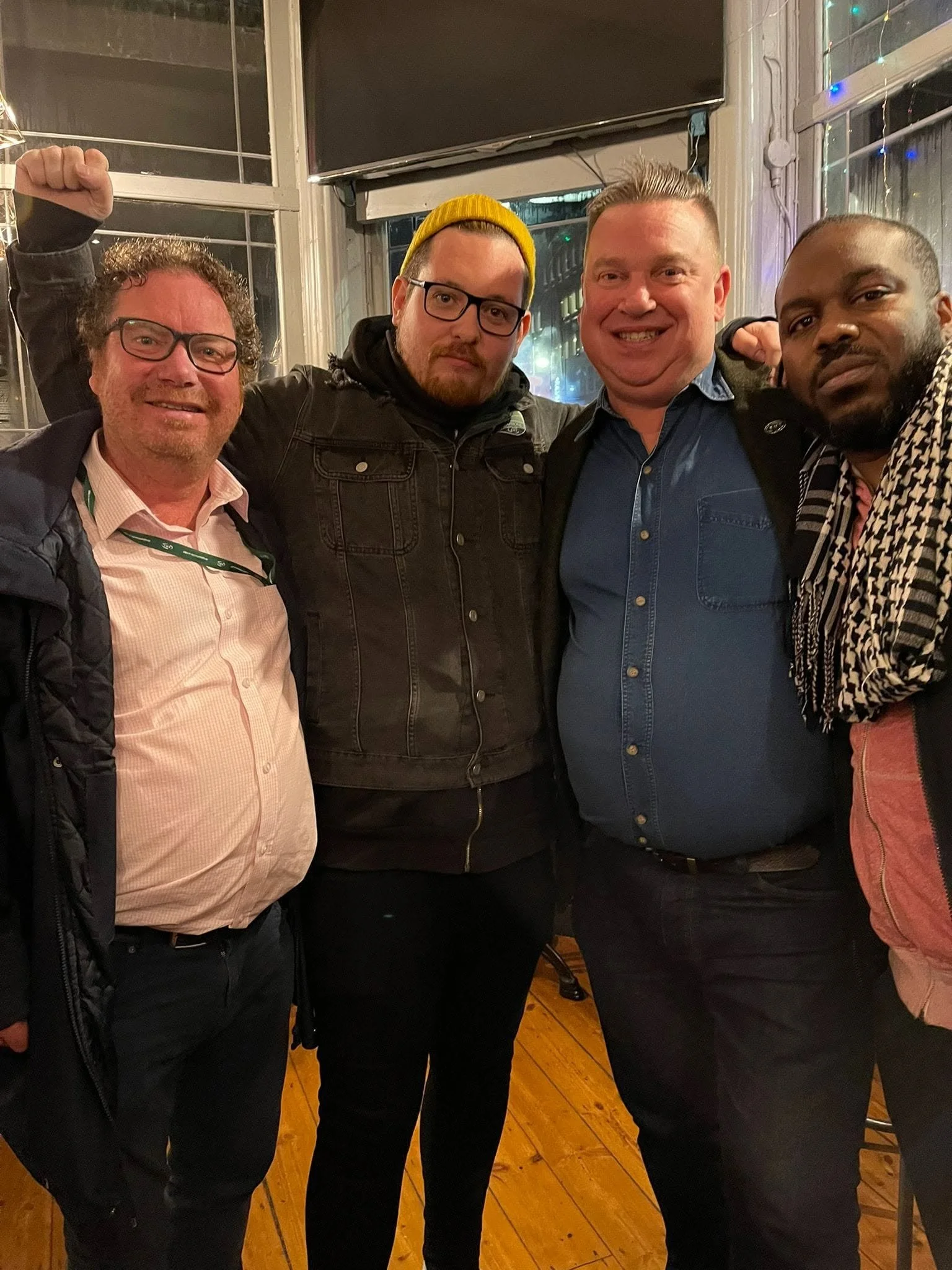 Four men posing together indoors at night, smiling and showing enthusiasm, with one man raising his fist.