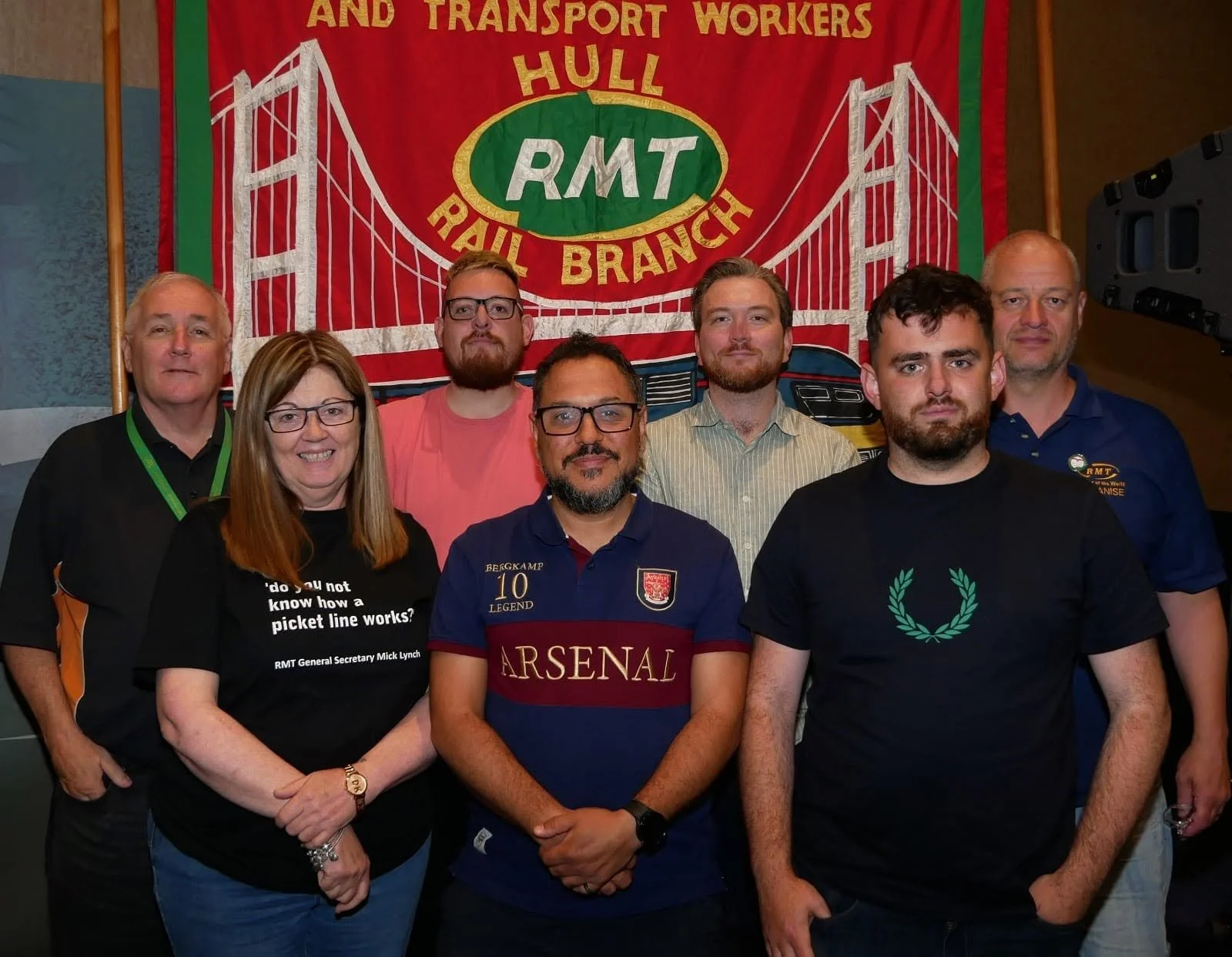 Group of seven people standing in front of a red banner with a bridge illustration and the text 'AM AND TRANSPORT WORKERS HULL RMT RAIL BRANCH'. The group includes four men and three women, some wearing shirts with logos or text, and they are posing for a photo indoors.