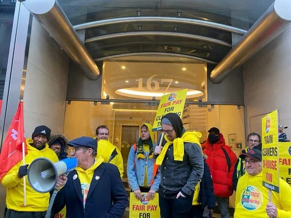 Group of people protesting outside a building, holding signs that read 'Fair Pay for Cleaners' and 'Justice for Cleaners.' Some are wearing yellow jackets and hats, and one person is speaking into a megaphone.
