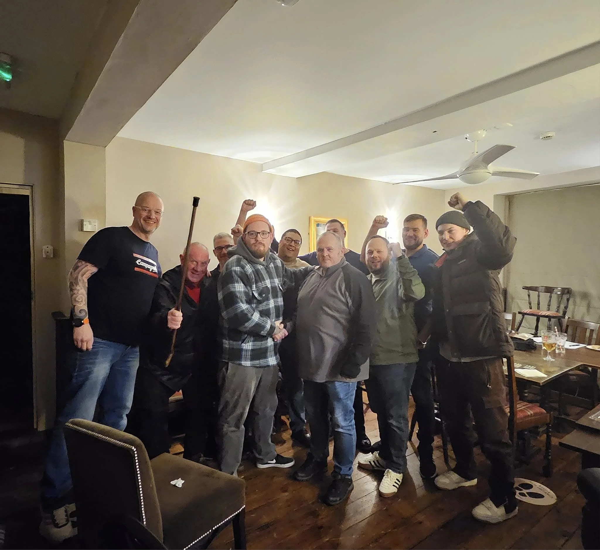 A group of men at a social gathering indoors, some with raised fists in celebration, smiling, and wearing casual clothing.