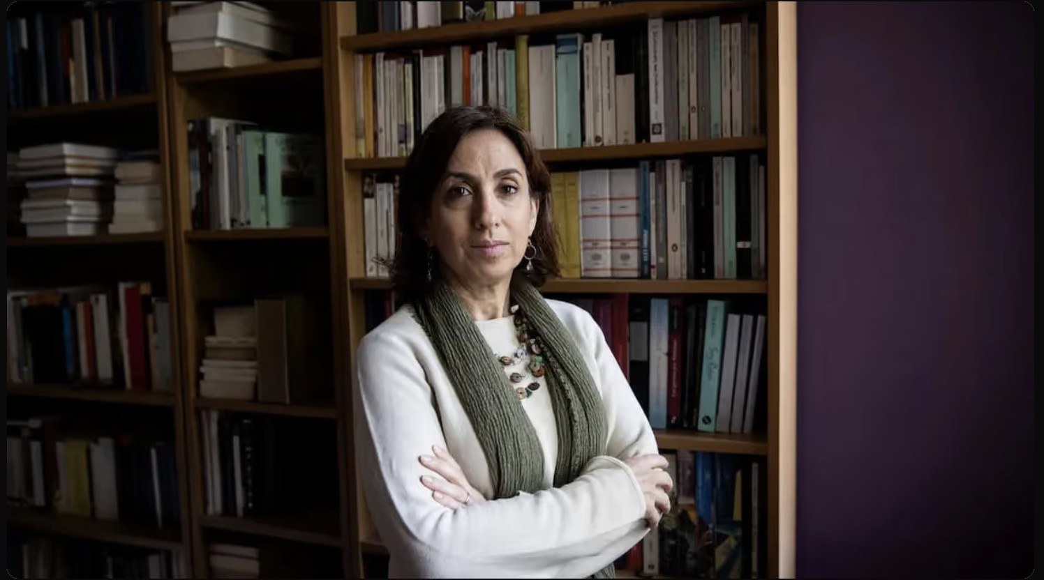 A woman with dark brown hair, wearing a white sweater, a green scarf, and a necklace, standing with crossed arms in front of a wooden bookshelf filled with books.