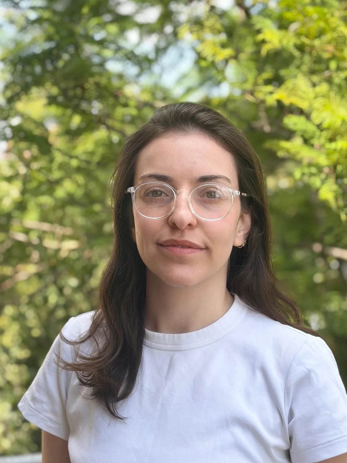 A young woman with long dark hair, clear glasses, and a white T-shirt standing outdoors, with green tree leaves in the background.
